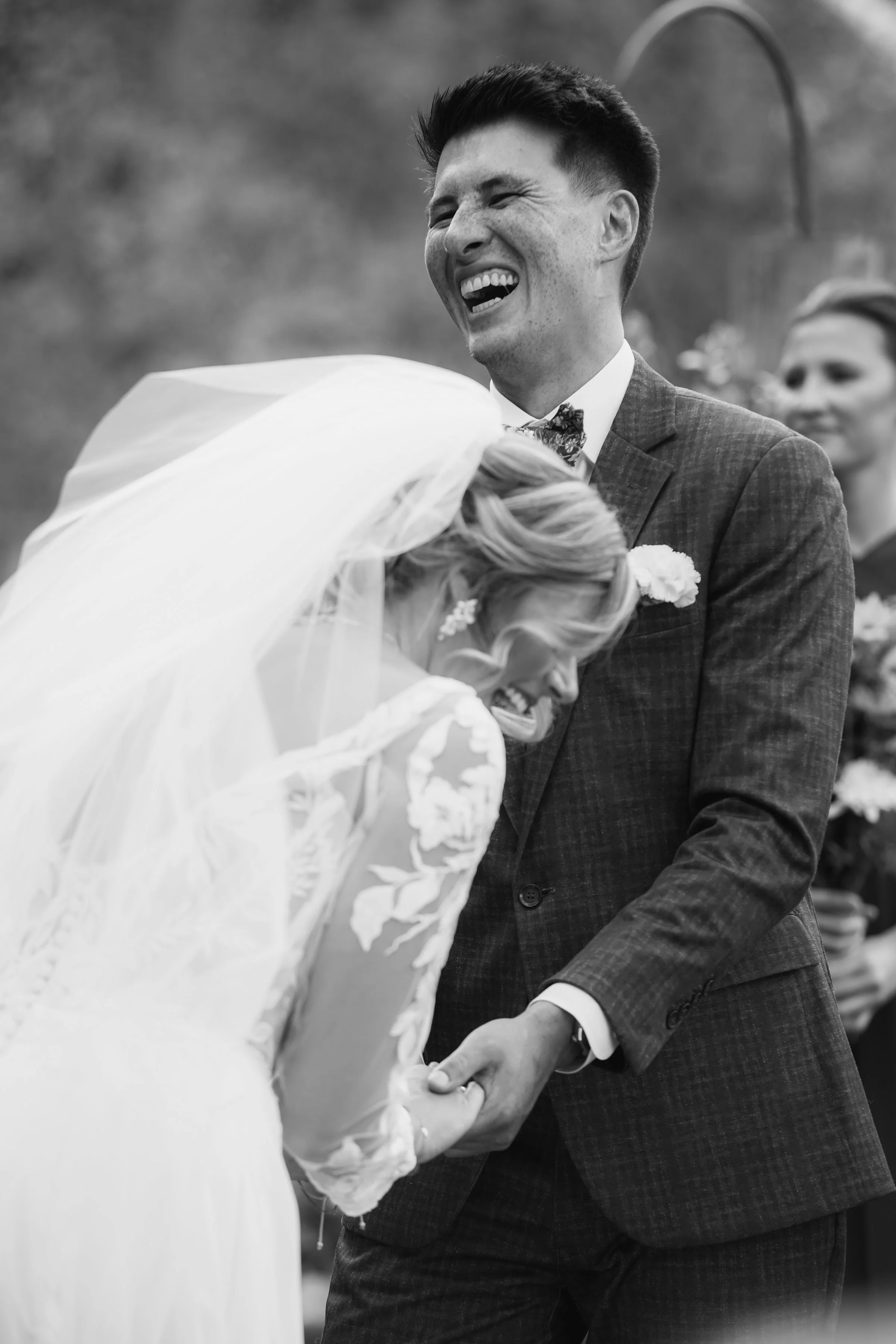 A black and white photo of a happy wedding couple, with the groom smiling while holding hands with the bride, who is wearing a wedding dress and veil.