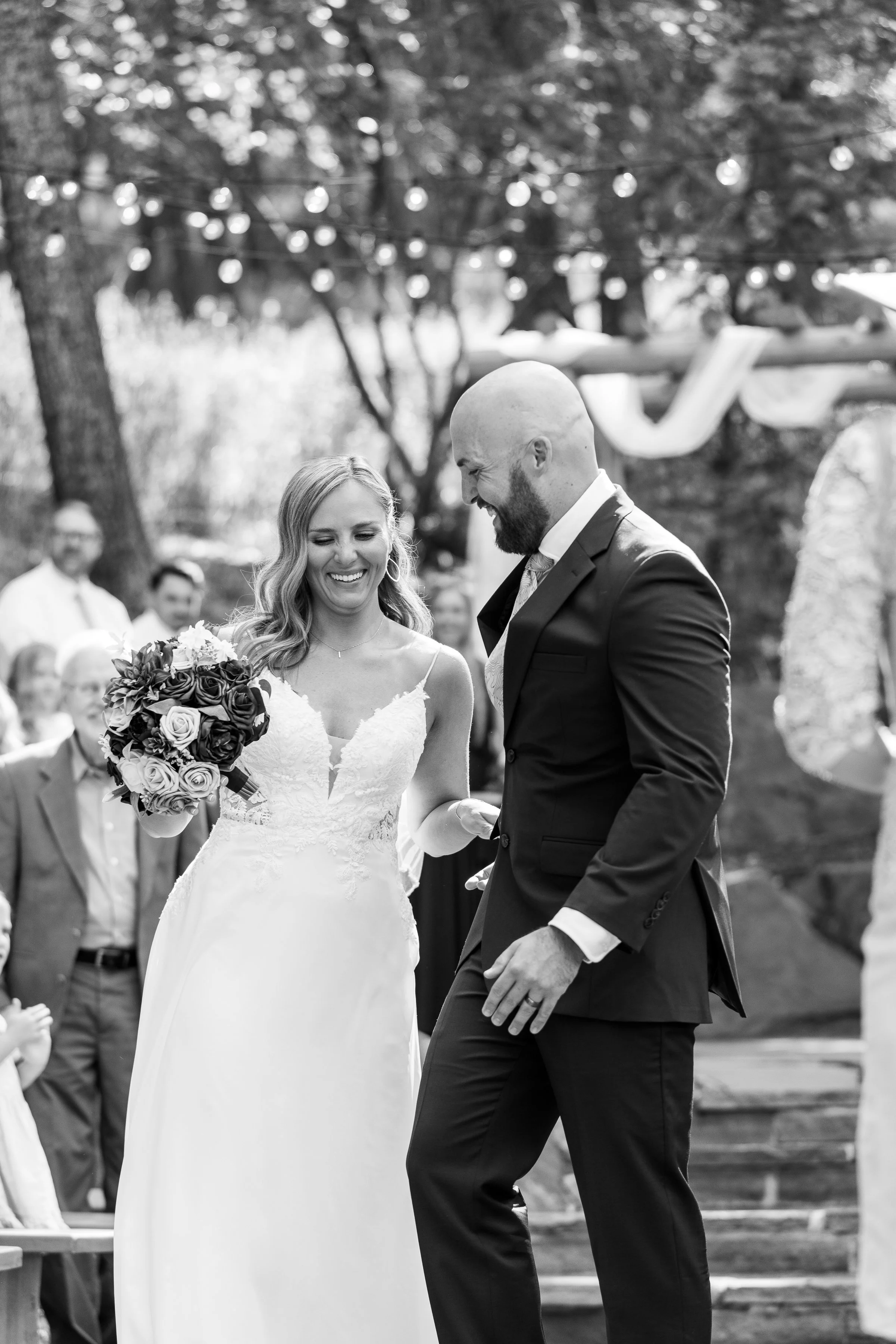 A bride and groom smiling at each other during their outdoor wedding ceremony, with guests in the background.