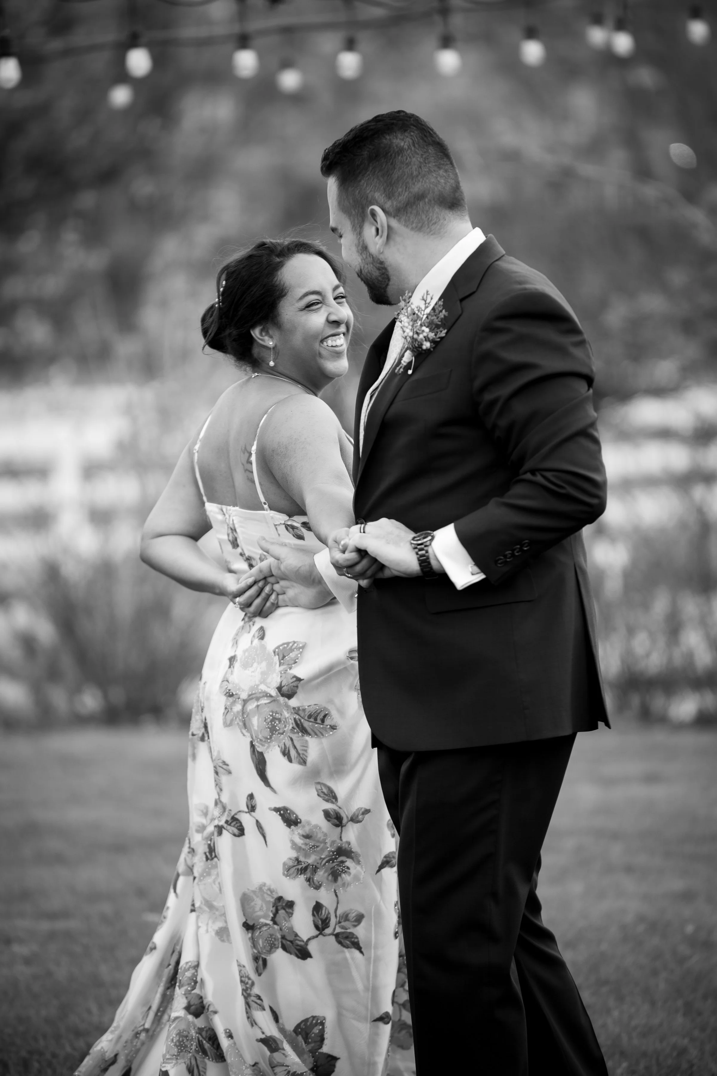 A black and white photo of a smiling woman in a floral dress and a man in a suit dancing outdoors, holding hands and embracing.