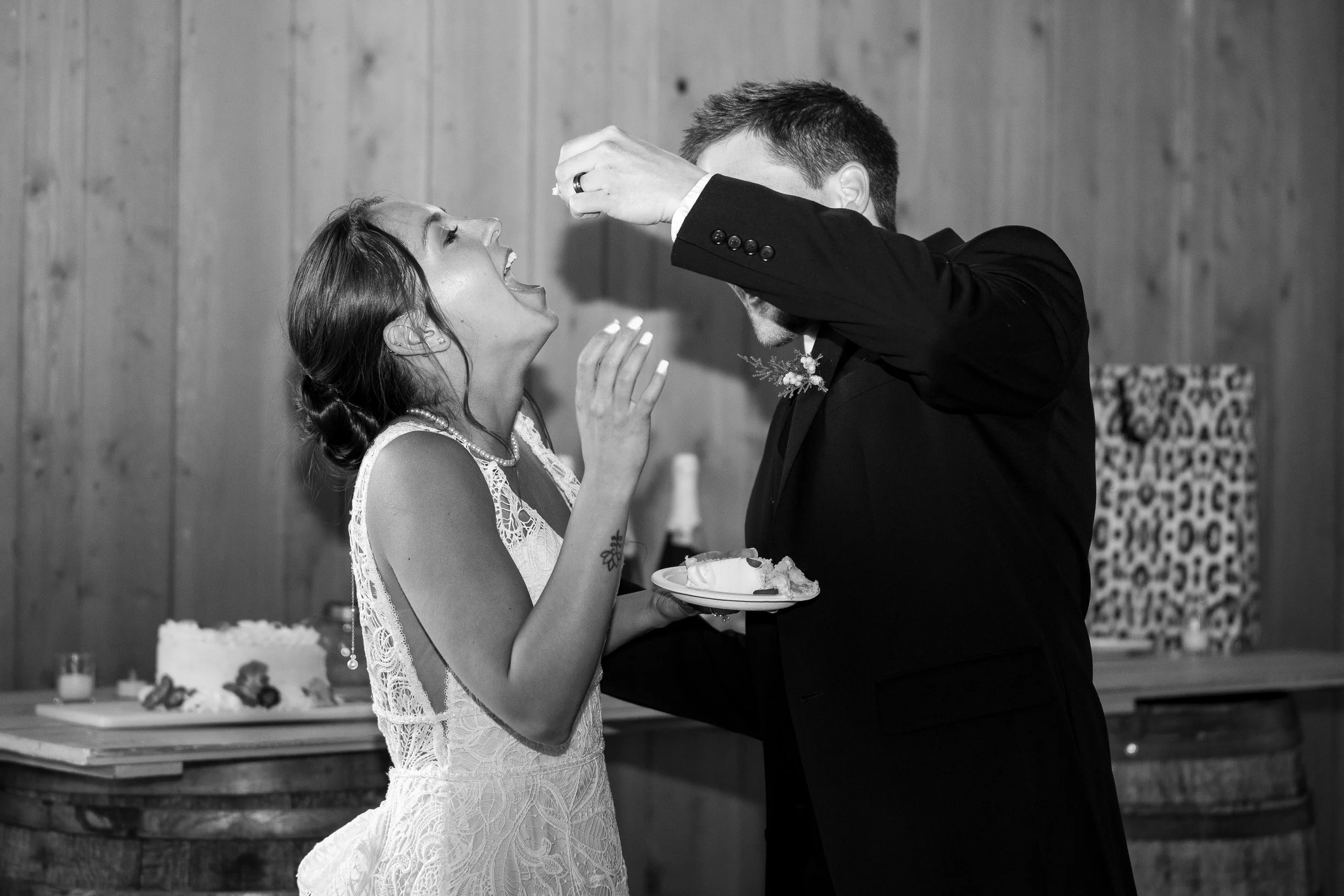 Black and white photo of a bride and groom at their wedding reception, with the groom feeding the bride piece of cake, both smiling, and cake on a table in the background.