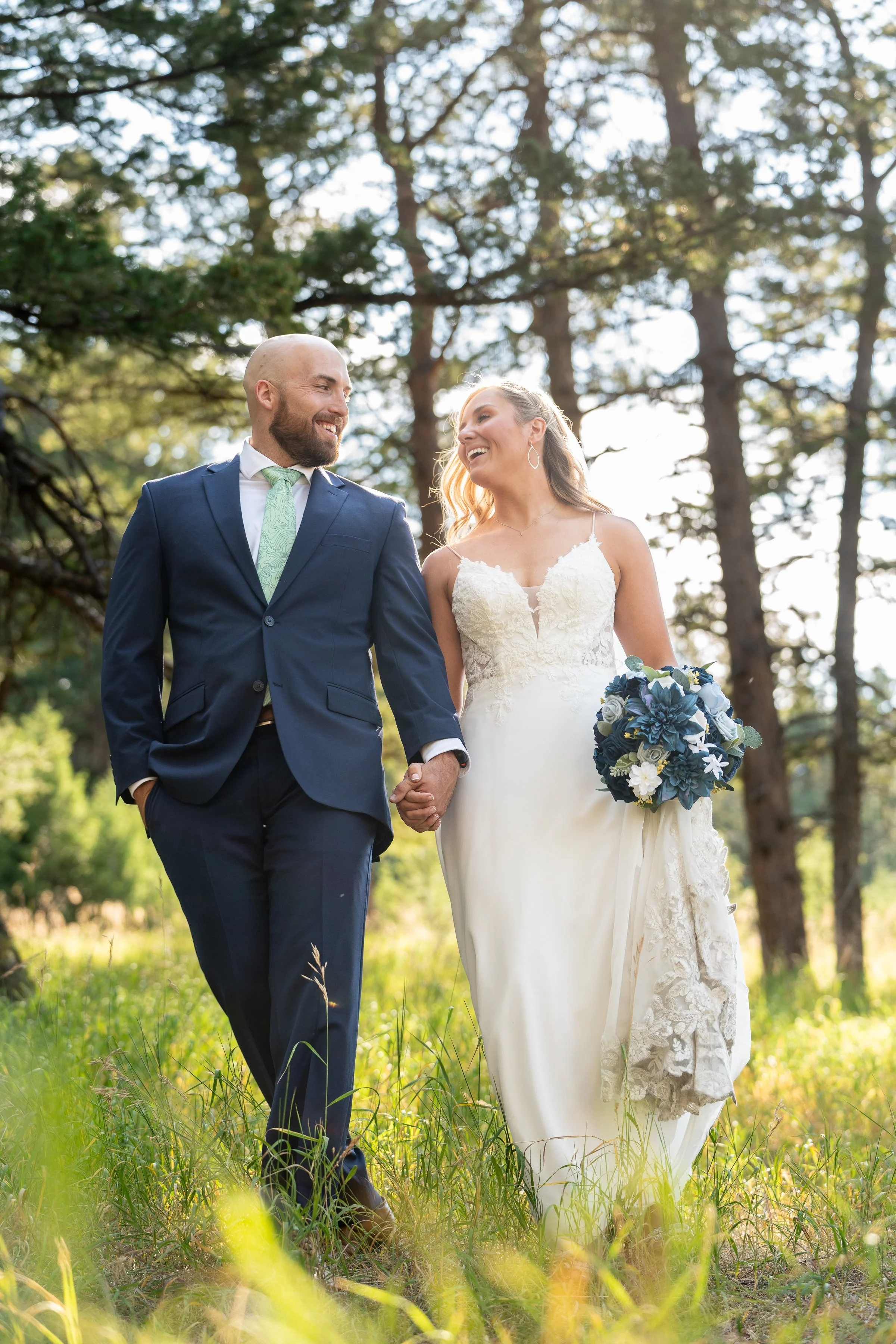 A newlywed couple walking hand-in-hand outdoors in a forest, smiling at each other. The groom is wearing a blue suit with a light green tie, and the bride is in a white wedding dress holding a bouquet of blue and white flowers.