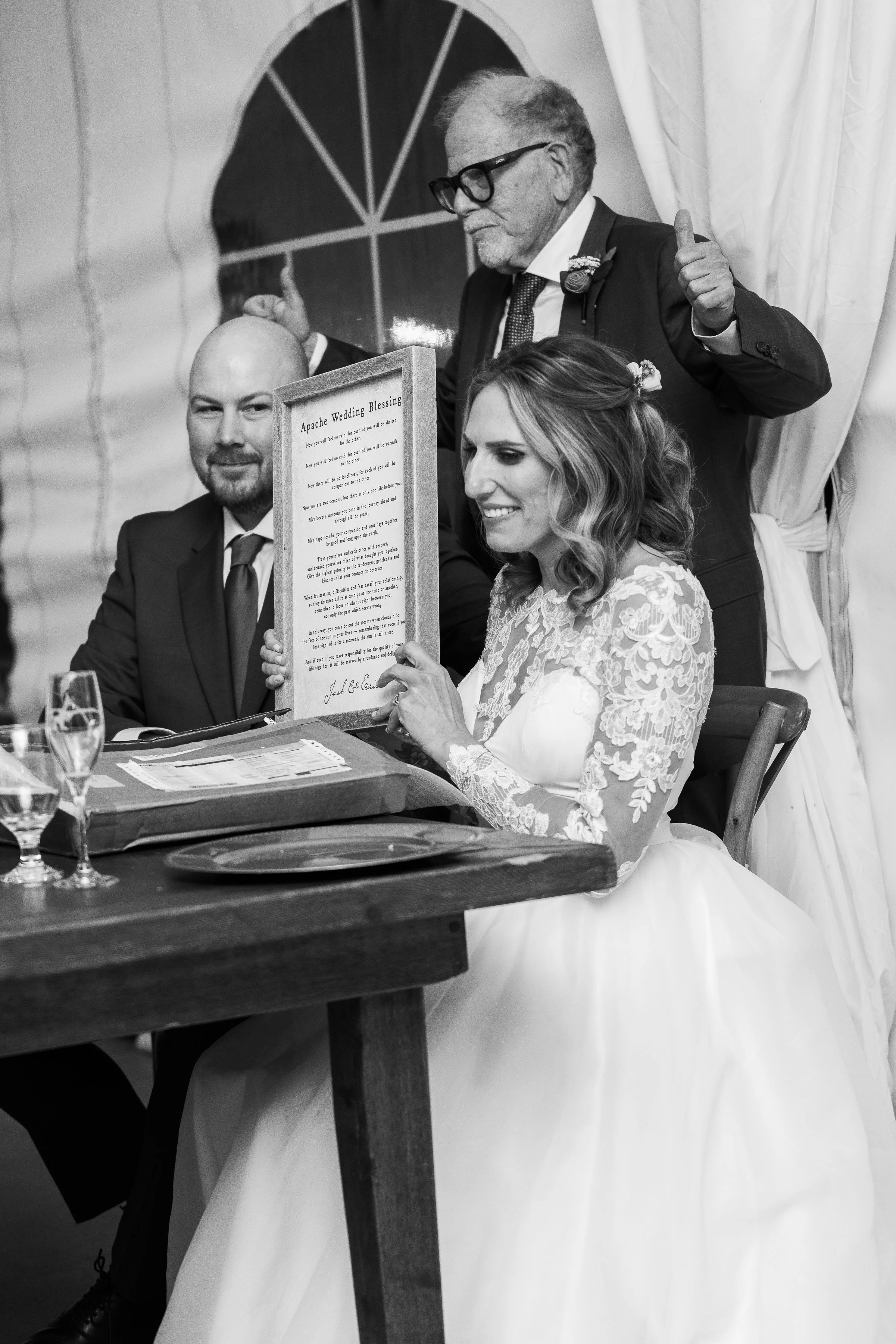 A bride in a lace wedding dress reads a document titled 'Apache Wedding Blessing' during a wedding ceremony, with a man in a suit and another man with glasses behind her, giving a thumbs-up.