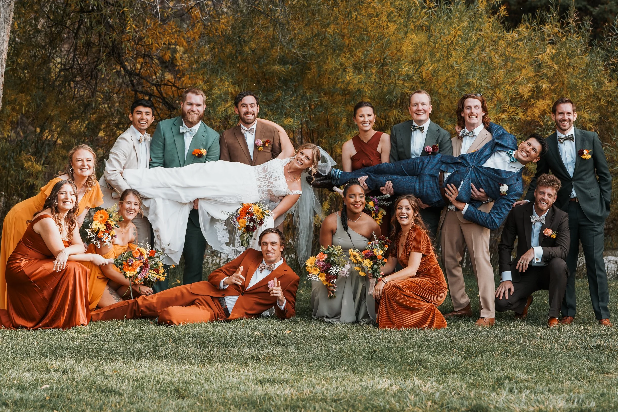A group of people at a wedding, with two individuals being lifted horizontally, one woman in a white wedding dress and one man in a blue plaid suit, among friends and family outdoors with fall foliage in the background.