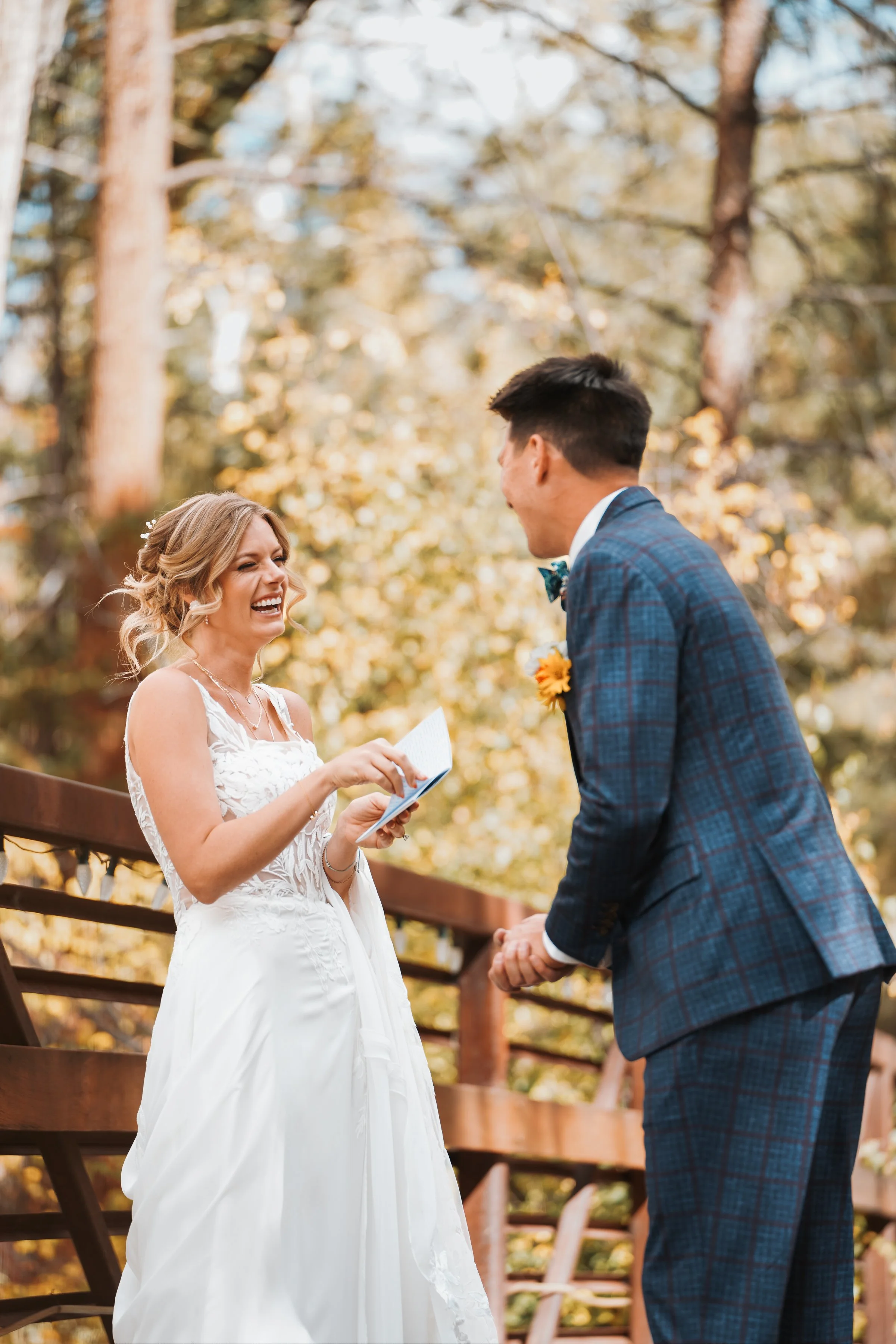A woman in a white wedding dress laughs as she reads her vows to a man in a blue plaid suit on an outdoor wooden bridge during a wedding ceremony, with trees in the background.