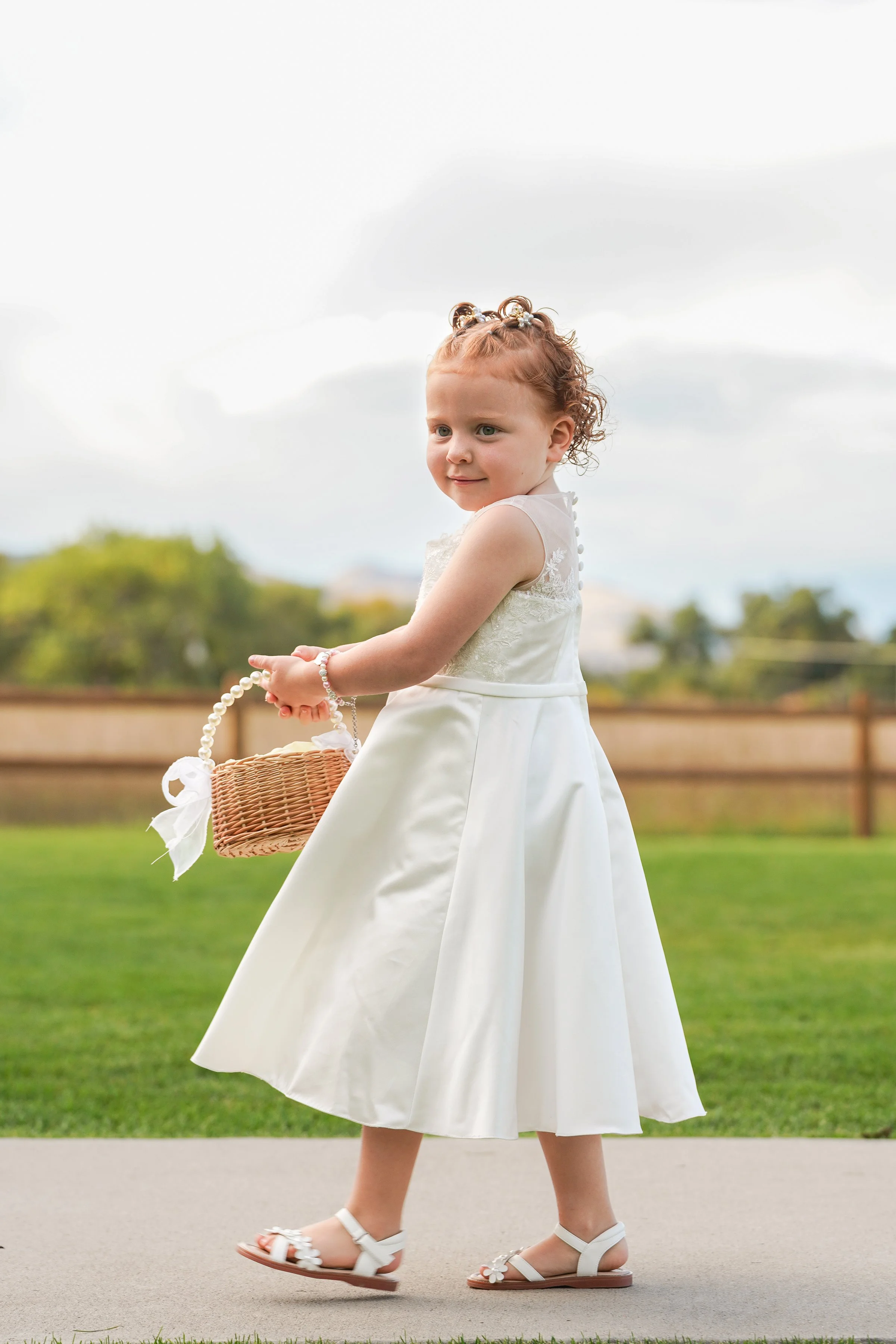 A young girl in a white dress walking outdoors on a sidewalk, holding a small wicker basket, with a grassy yard and cloudy sky in the background.