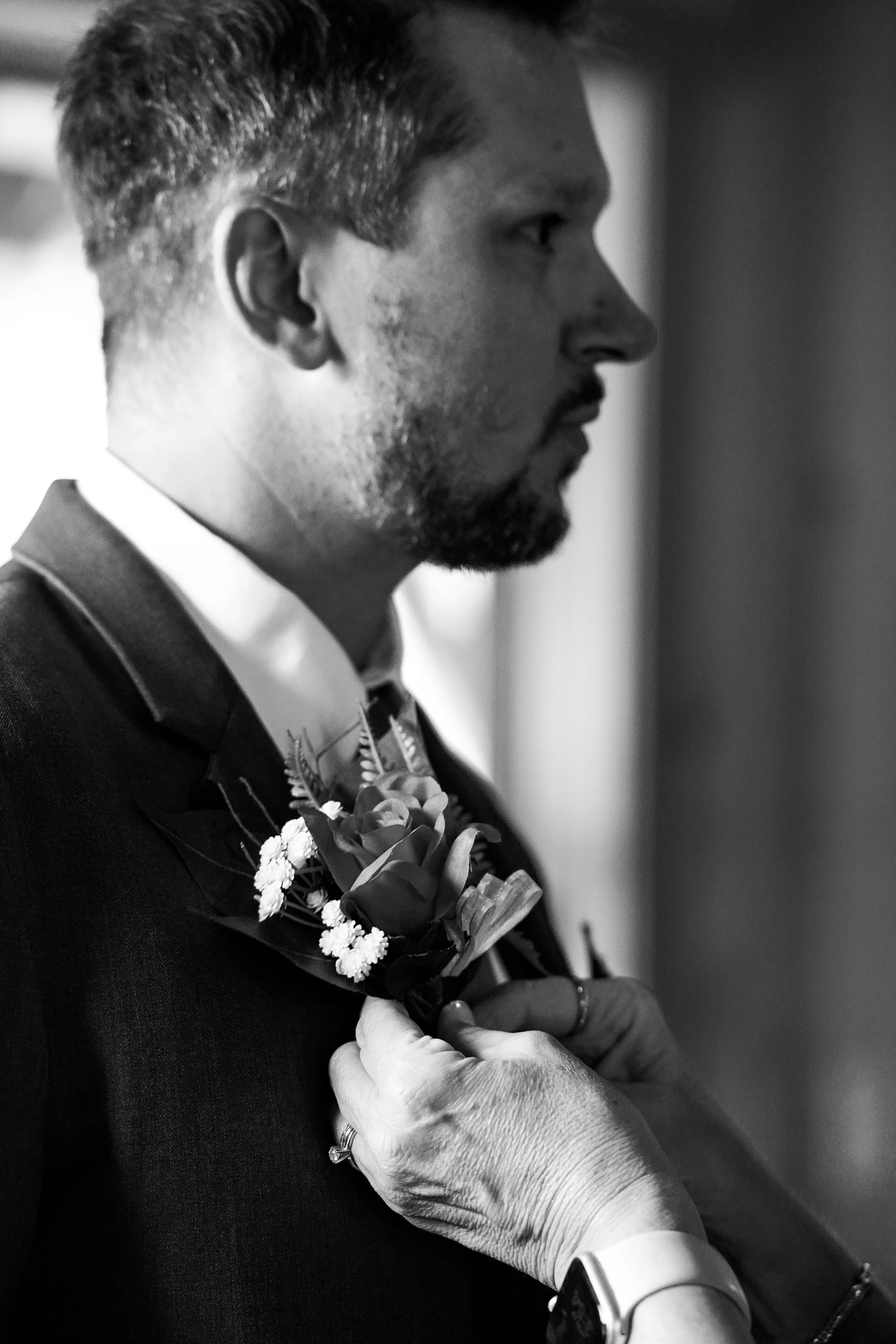 A man in a suit is having a boutonniere pinned on his lapel by a person with a ring and a watch. The image is in black and white.