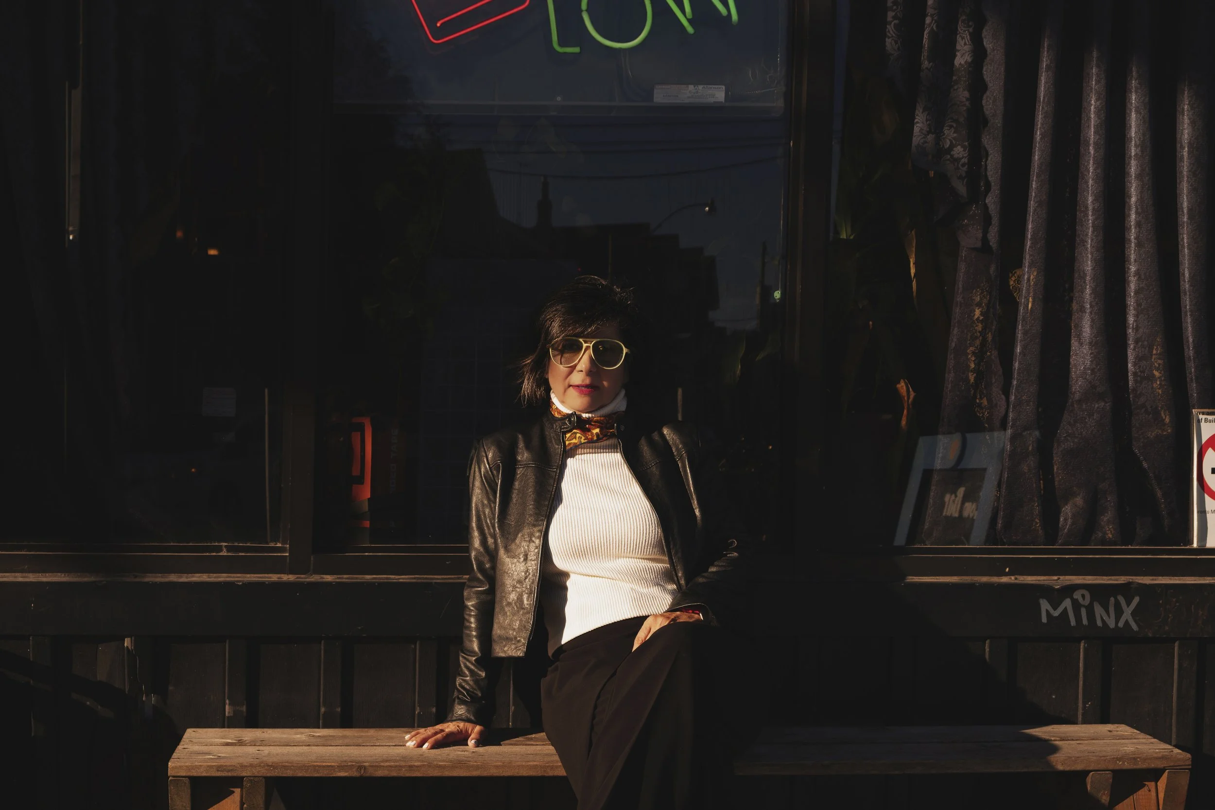 A woman with short dark hair and yellow sunglasses sitting on a wooden bench outside a storefront with dark curtains and a neon sign.