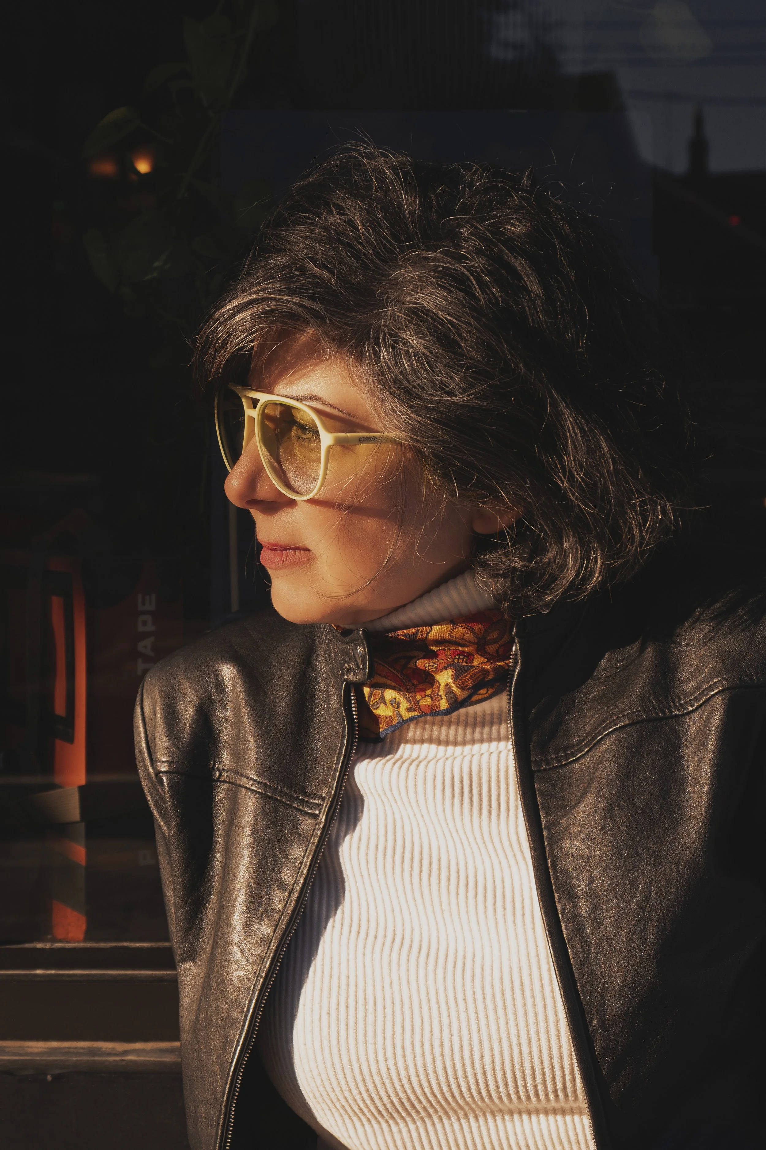 A woman with brown, wavy hair wearing yellow sunglasses, a leather jacket, a patterned scarf, and a cream ribbed shirt, looking to her left in sunlight.