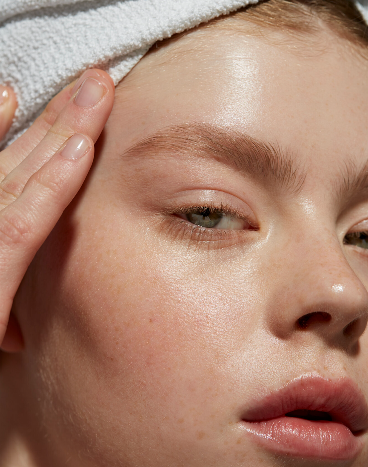 Close-up of a woman's face with a towel wrapped around her head, gently touching her forehead with her fingers, showing clear skin with minimal makeup.