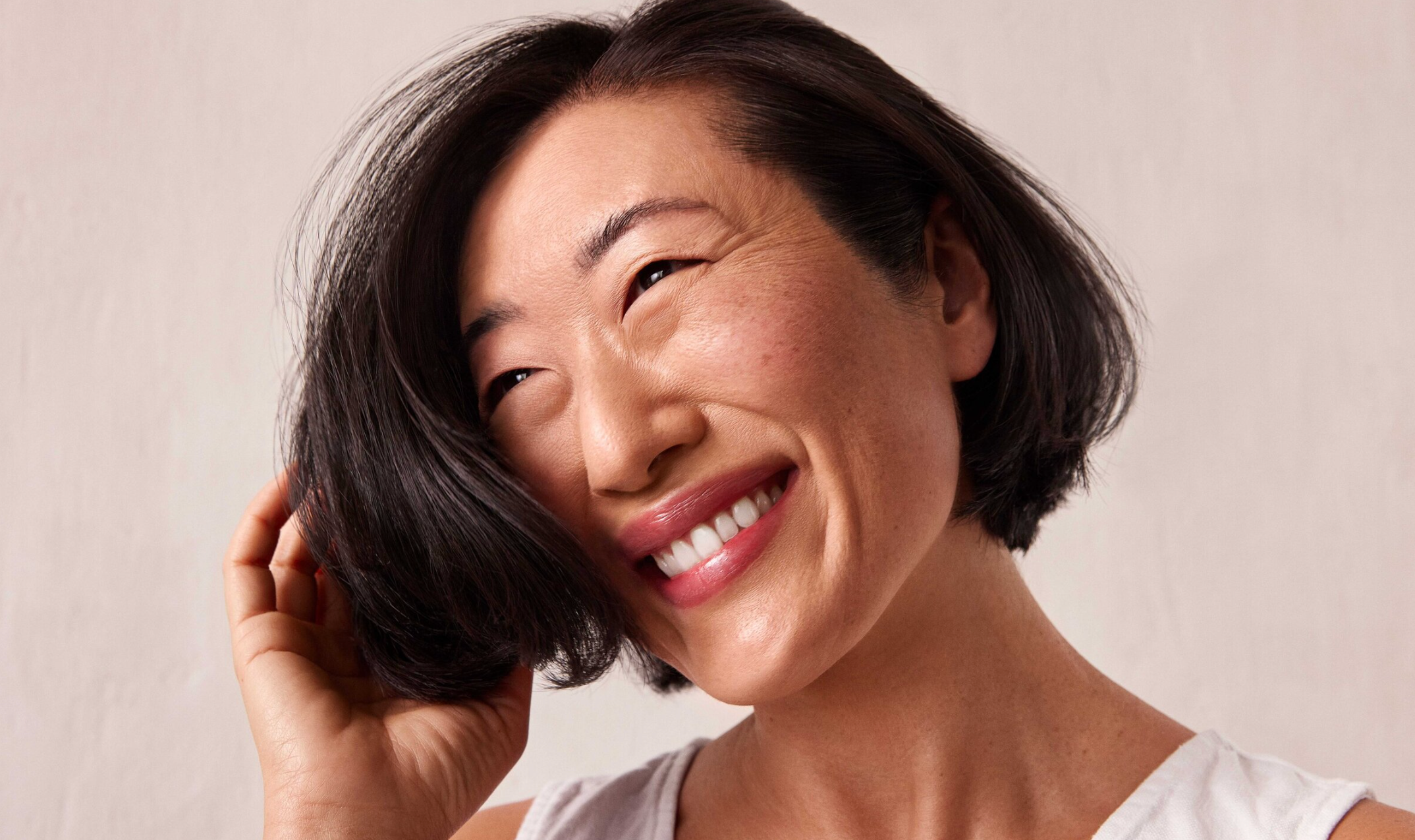 Close-up of a smiling woman with short black hair, touching her hair, against a light plain background.