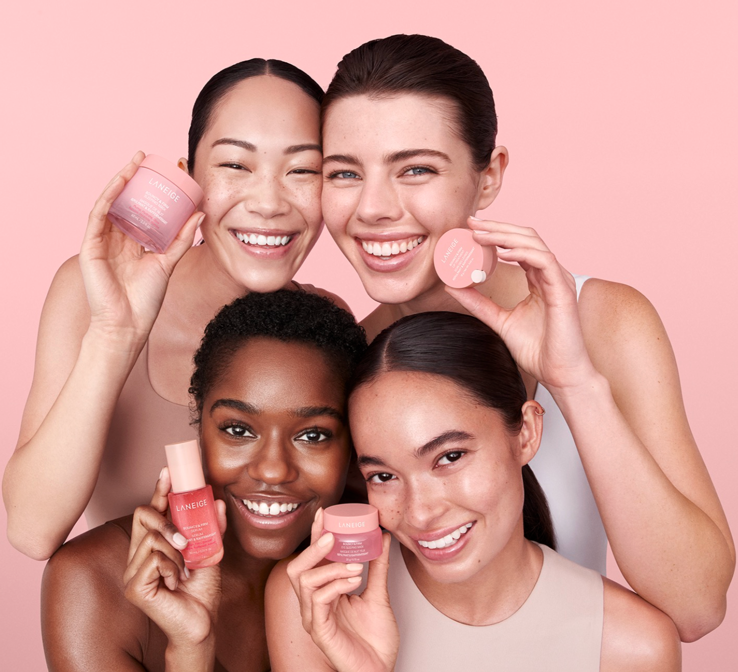 Four women smiling and holding pink skincare products against a pink background.