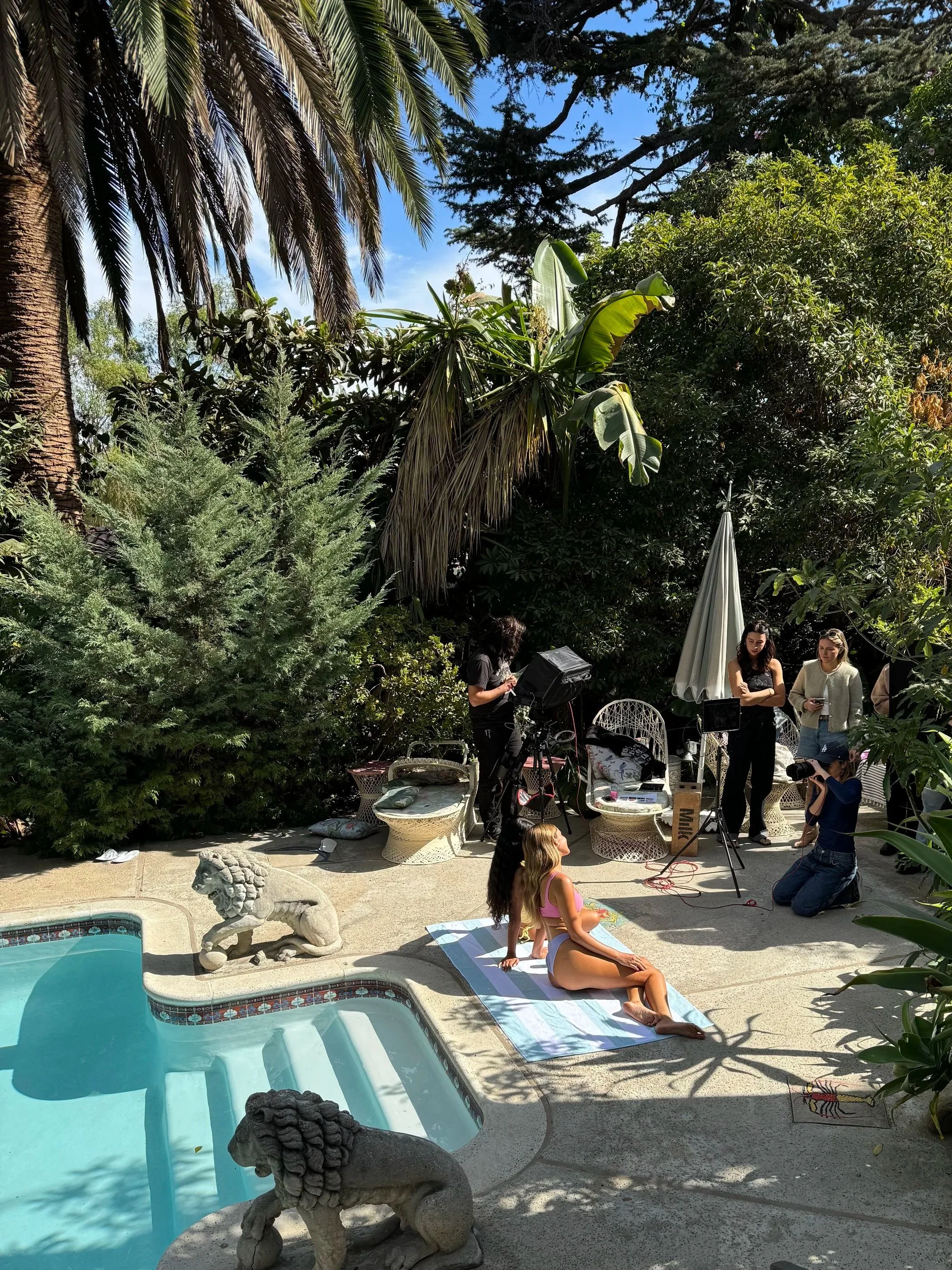 A woman in a swimsuit poses on a towel by a swimming pool as a film crew sets up equipment outdoors surrounded by lush green trees and plants, with some people working and taking photos.