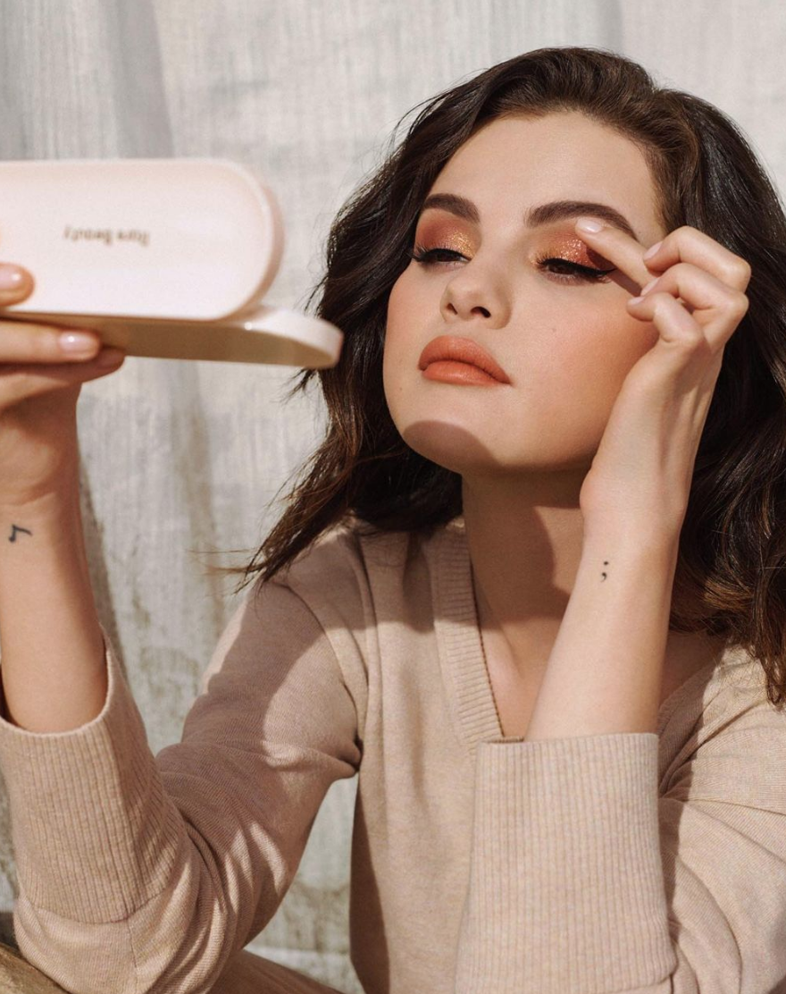 Selena Gomez is applying makeup while looking into a mirror. She is wearing a beige long-sleeve top and is holding a small mirror in her left hand.