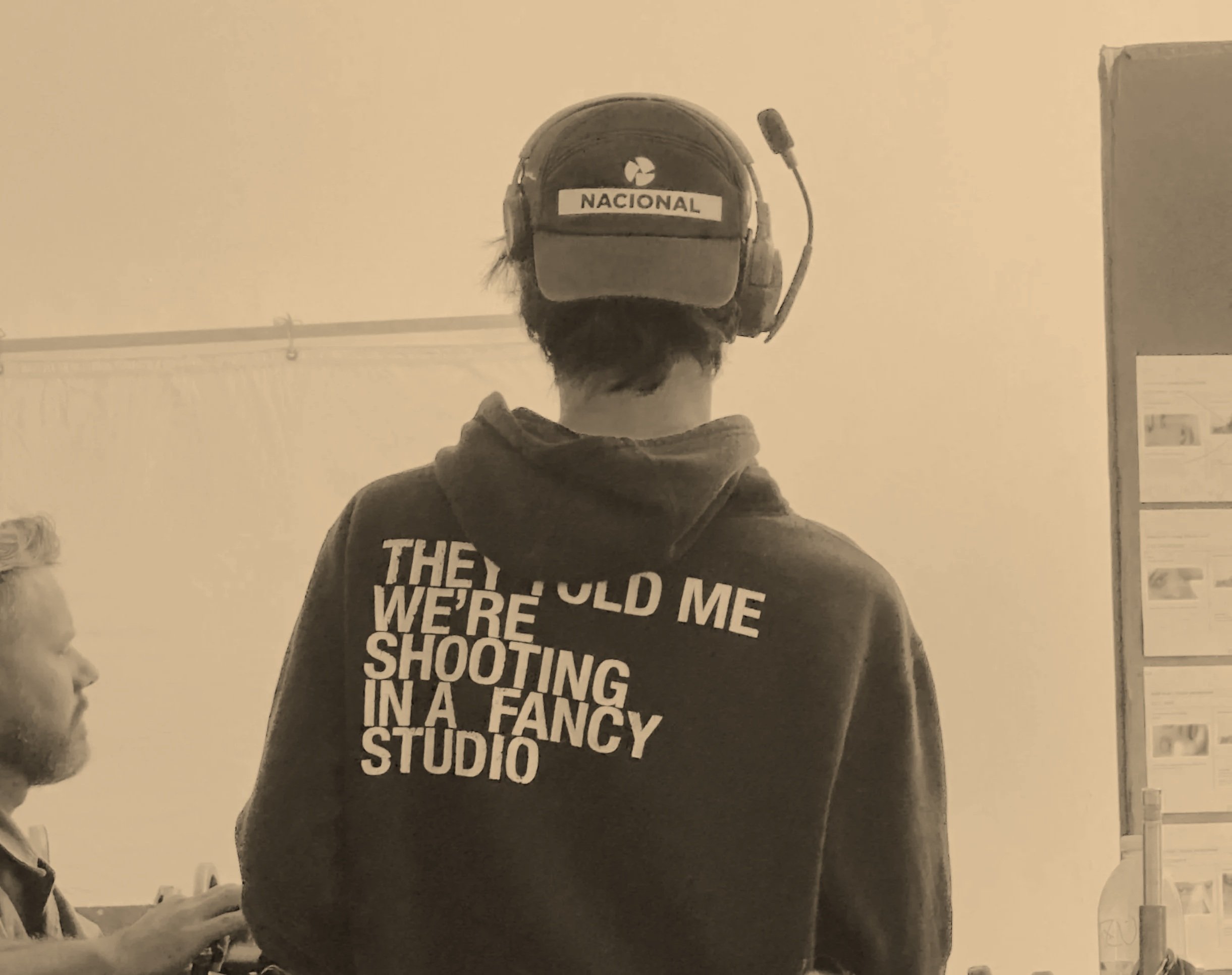 Person wearing a headset and hoodie that reads 'they told me we're shooting in a fancy studio', viewed from behind, in a studio setting, with another person visible in the background.