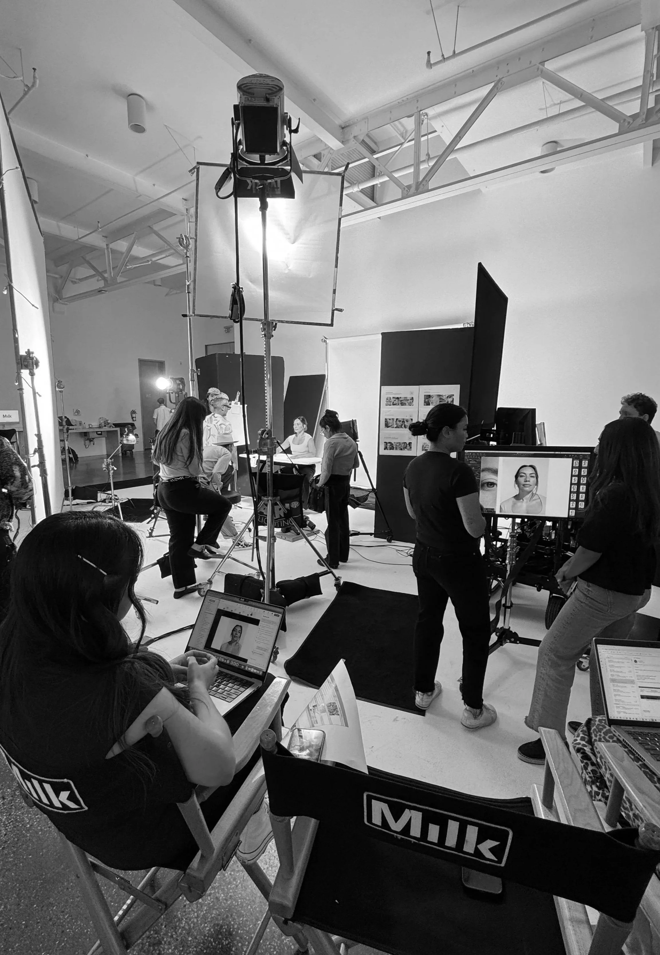 Behind-the-scenes of a photo shoot with makeup artists and photographers, equipment, and a model on a white background.