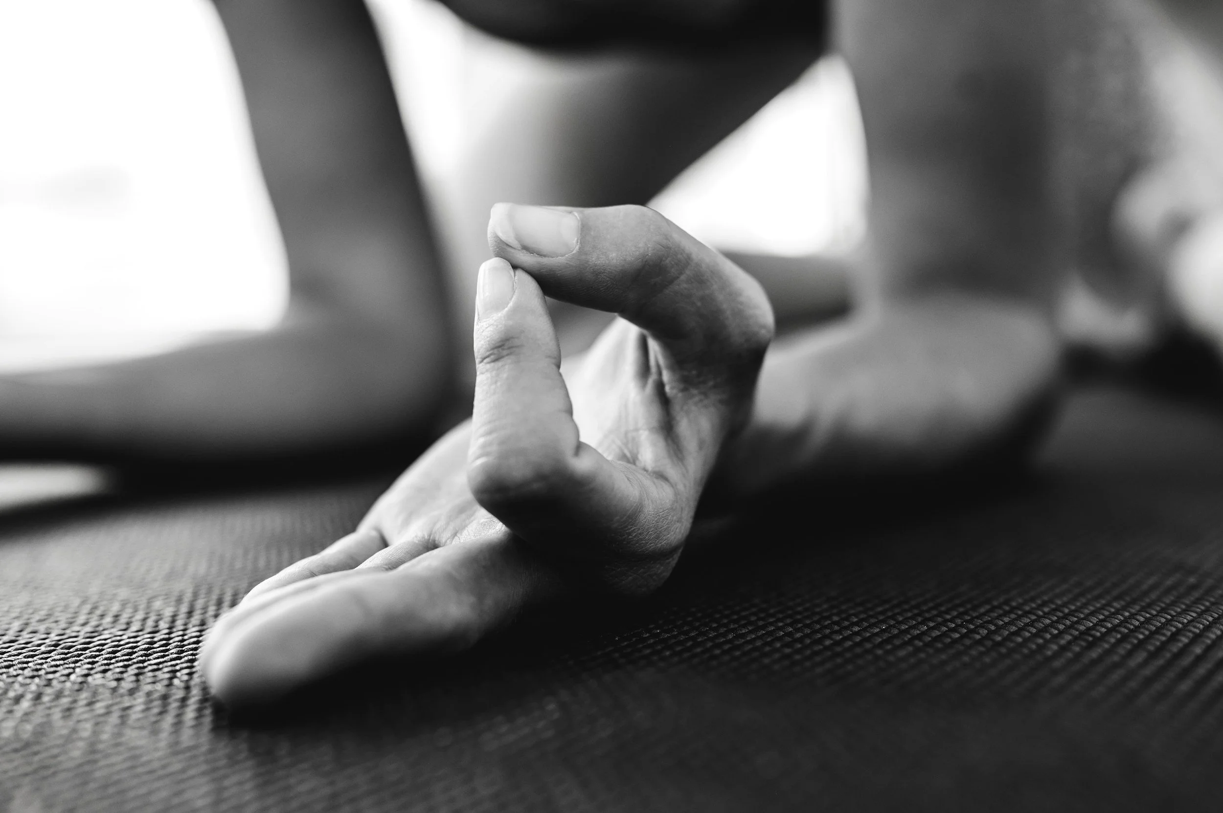 Close-up black and white photo of a person doing yoga or stretching on a textured mat, focusing on their hand reaching out with fingers slightly curled.