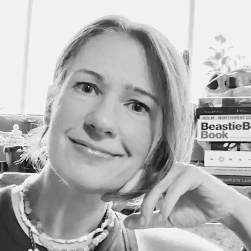 A woman with shoulder-length hair smiling, resting her head on her hand, near a stack of books including a 'Beastie B' book, a plant, and a window with natural light.