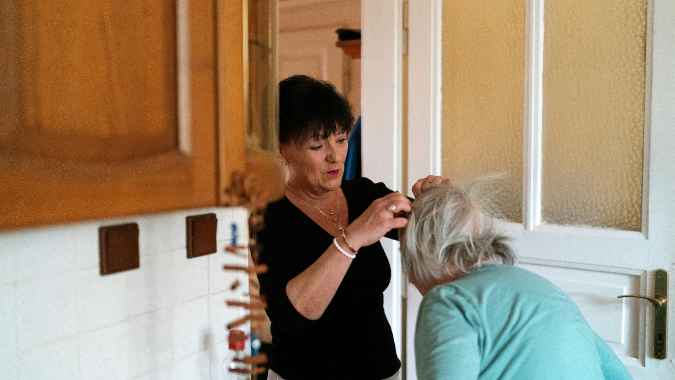 A woman with short dark hair helping an elderly woman with long gray hair put on earrings in a doorway.