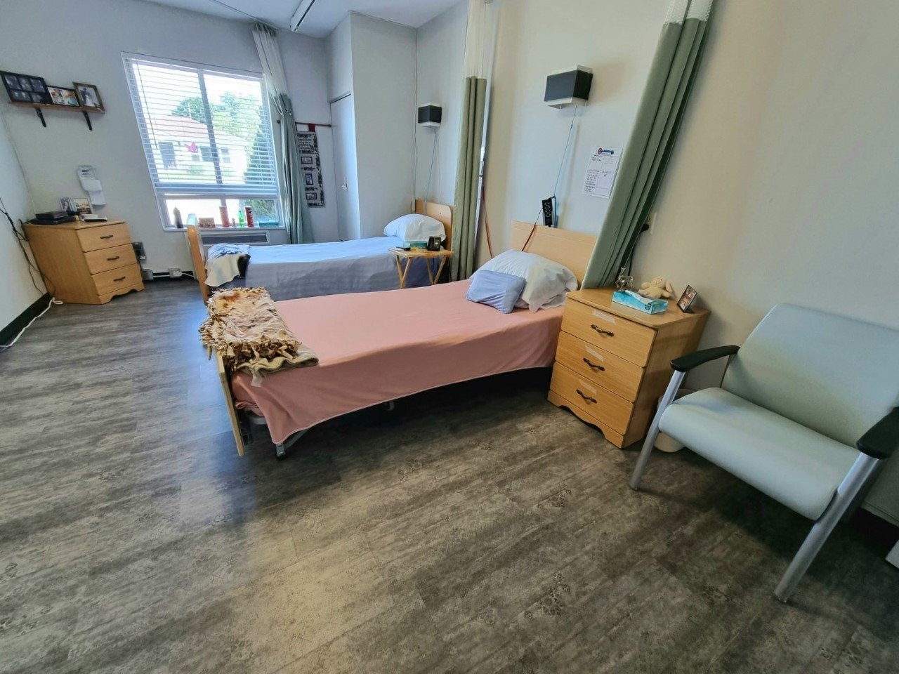A hospital room with two beds, one with pink sheets and one with gray sheets, each with pillows and blankets. There is a white chair with armrests next to a wooden nightstand, and another wooden dresser with a phone and photo frames. A window with bl