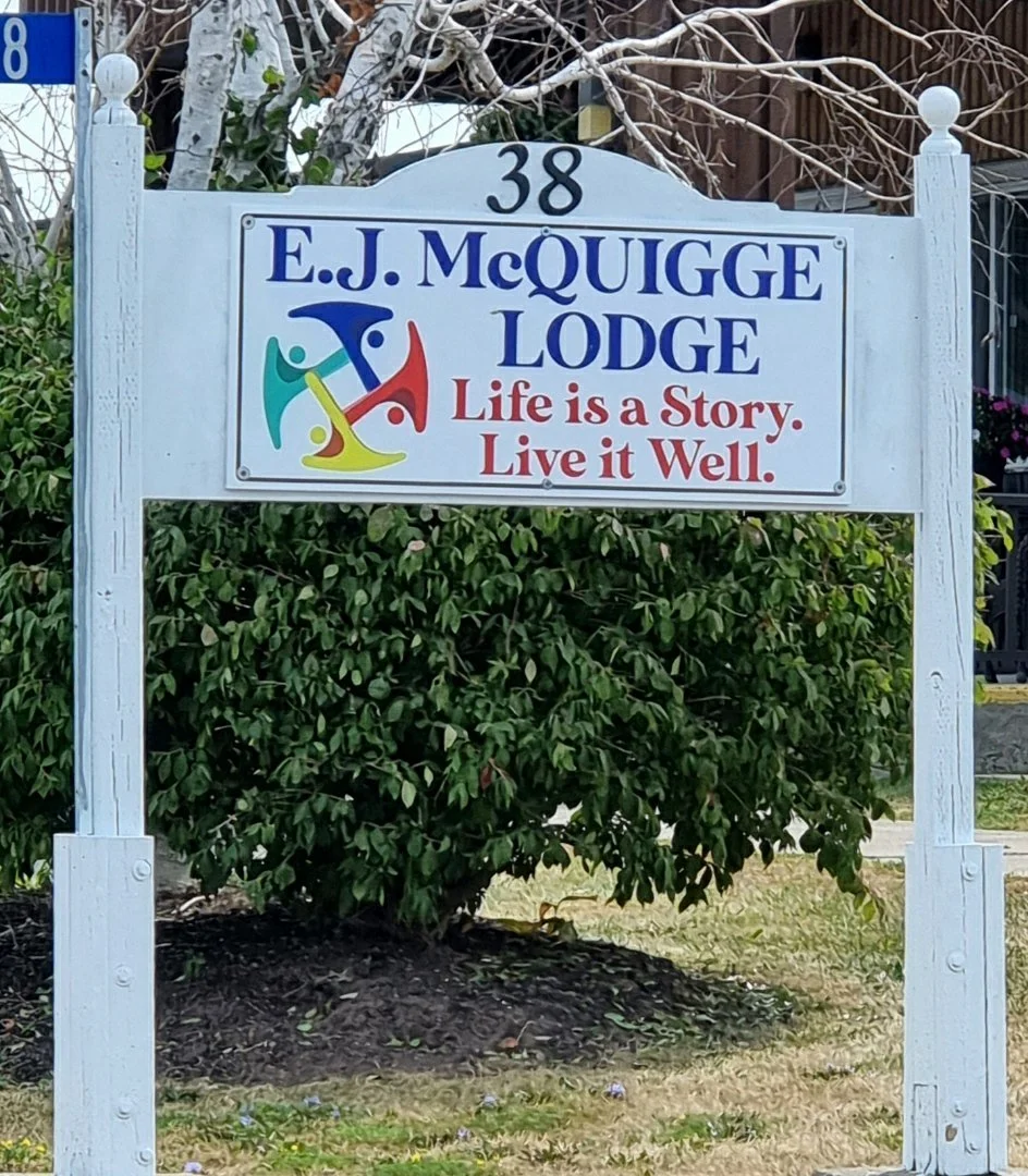 A white sign with the text '38 E.J. McQuigge Lodge' and a colorful logo, along with the message 'Life is a Story. Live it Well.' It is mounted on a white post in front of green bushes and some trees.
