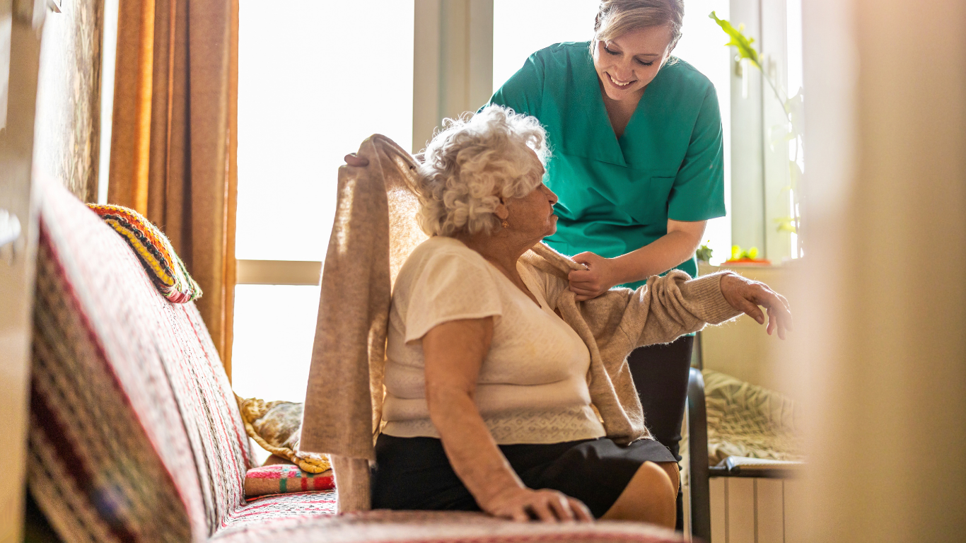 A caregiver helping an elderly woman get up from a chair in her home, with sunlight streaming in from a window.