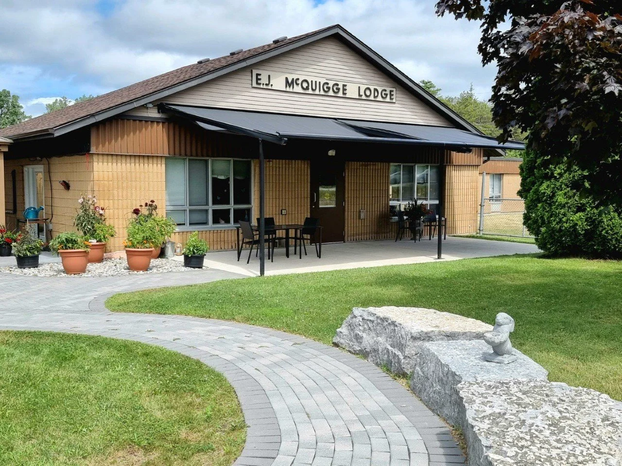 Exterior of E.J. McGuige Lodge with a brick walkway, flower pots, outdoor seating, and a stone duck sculpture in front of a grassy yard.