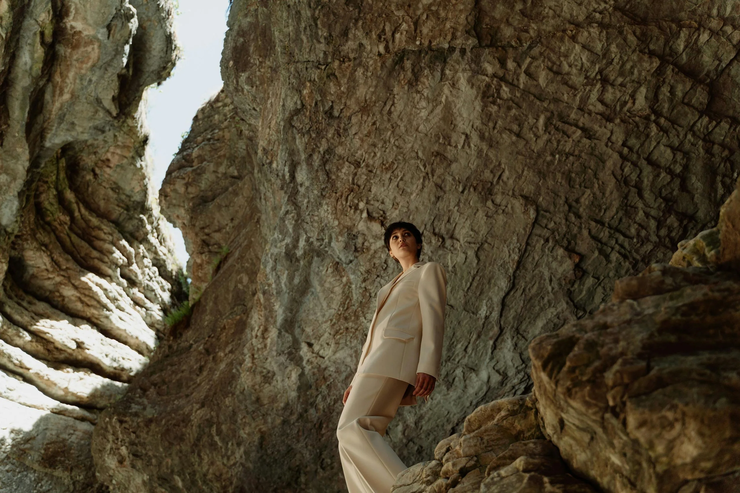 A woman in a beige suit standing among large rock formations and cliffs outside.