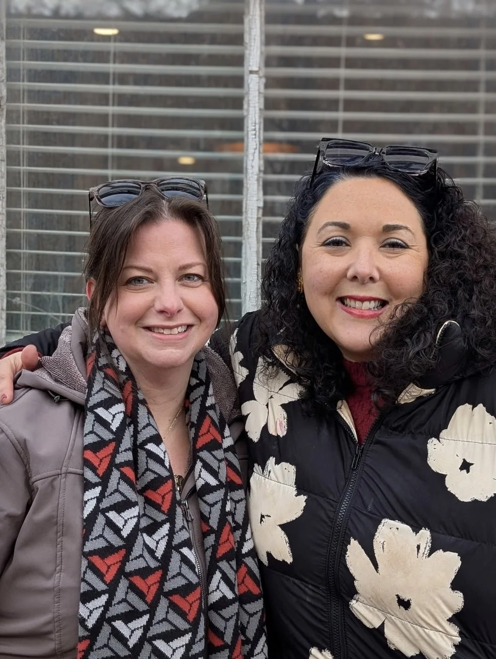 Two women smiling, standing outside in front of a large window with blinds. One woman has brown hair and is wearing a scarf with a geometric pattern and a jacket. The other woman has curly black hair, and is wearing glasses on her head, a floral-patterned jacket, and a red top.