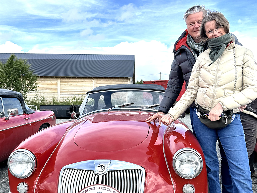 A couple standing next to a red vintage MG sports car outdoors, with a barn and other classic cars in the background.