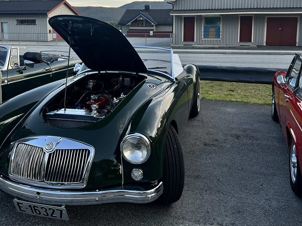 A vintage green MG sports car with the hood open, parked alongside other classic cars, on a street in a small town with houses and mountains in the background.