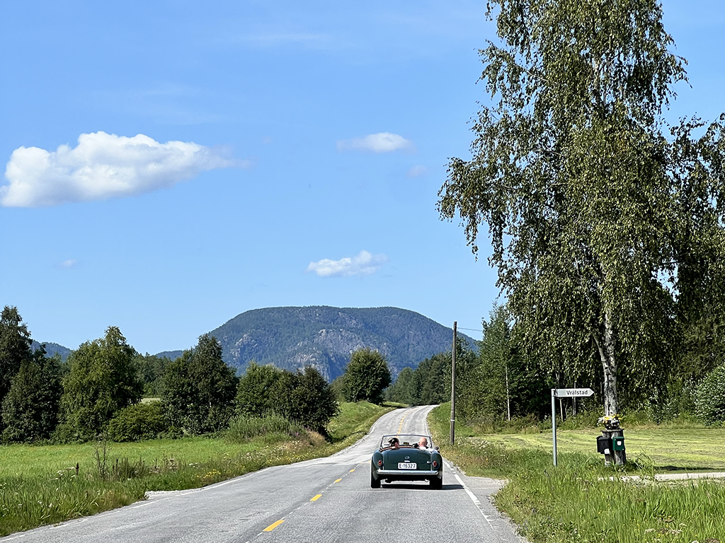 A rural road with a convertible car driving away, surrounded by green fields, trees, and a mountain in the background under a partly cloudy blue sky.
