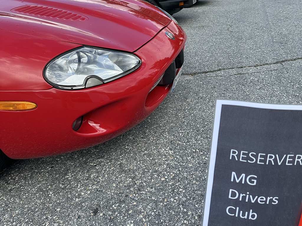 Close-up of the front of a red MG sports car parked on a paved surface with a sign in the foreground that reads "RESERVED MG Drivers Club".