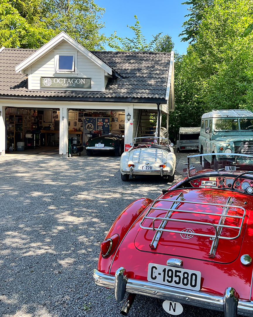 A collection of vintage cars parked outside a garage labeled 'MG Octagon Garage,' with trees and a bright blue sky in the background.