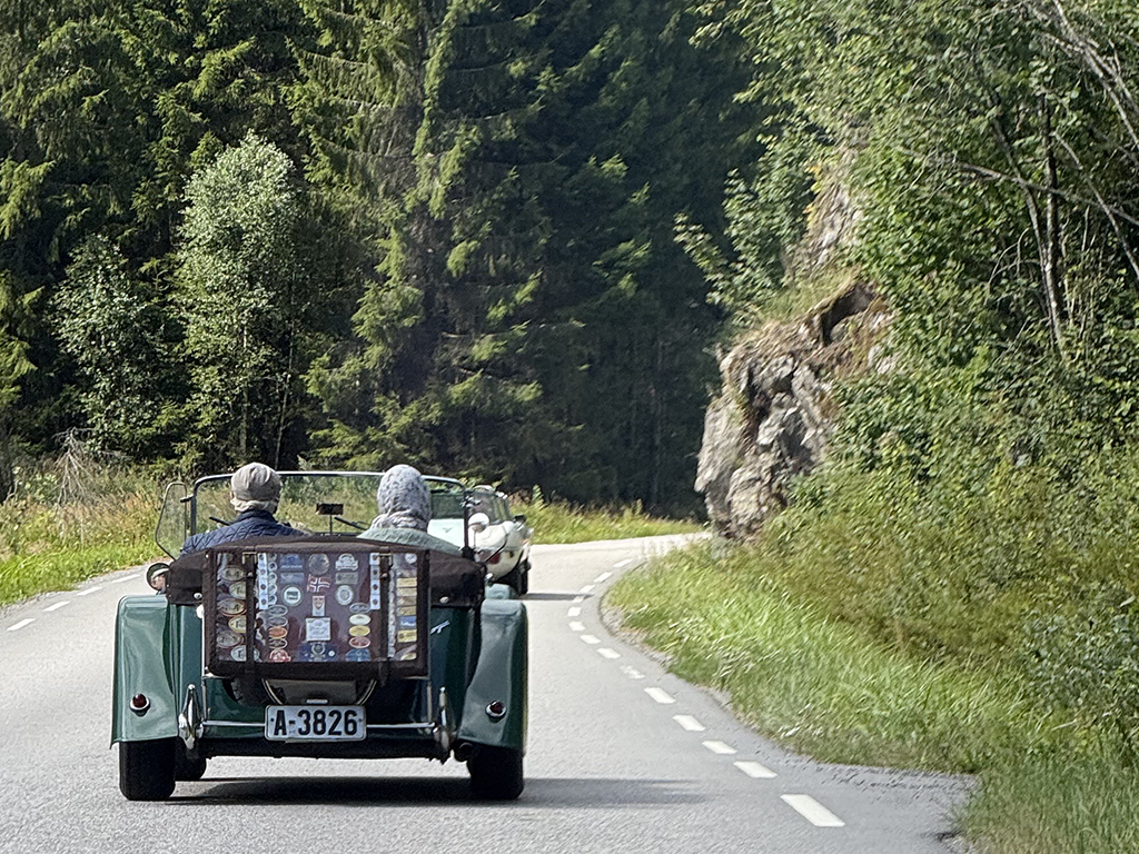 A couple driving in a vintage open-top car along a winding mountain road surrounded by trees and rocky cliffs.