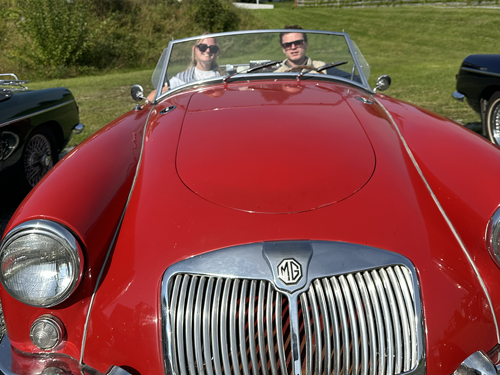 Red vintage MG convertible car with two people inside, a man and a woman, sitting on a grassy area during daytime.