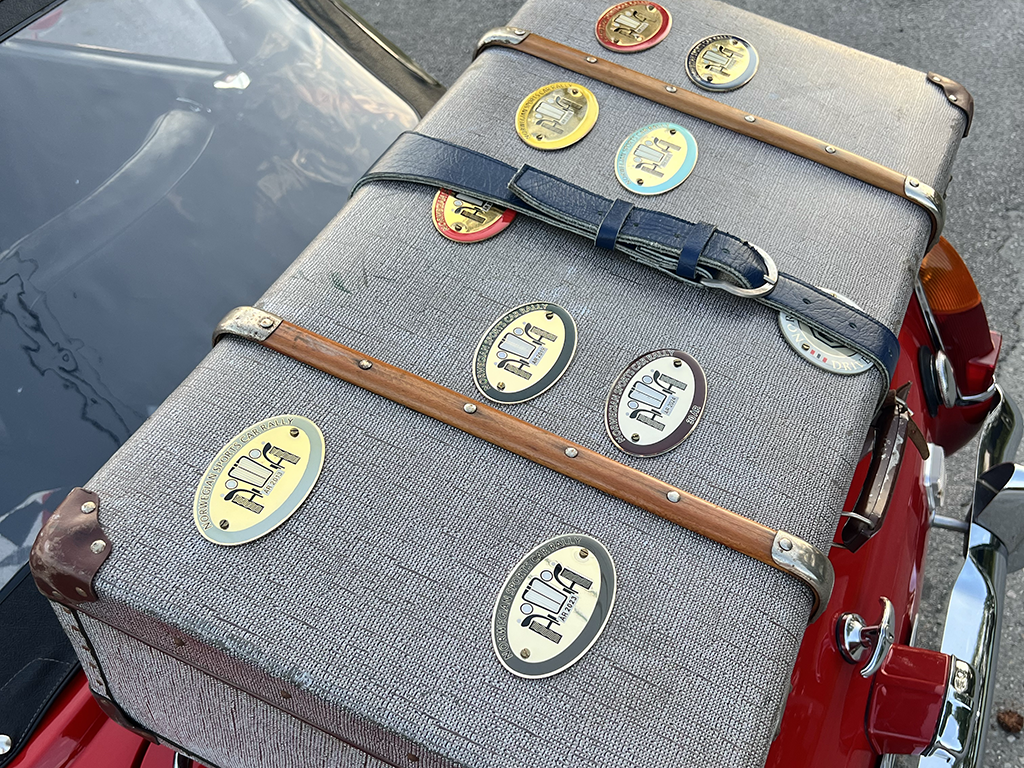 Two vintage suitcases with Travel stickers placed on the back of a red motorcycle, one suitcase has a black strap and the other has a brown strap, reflecting in a nearby car's windshield.