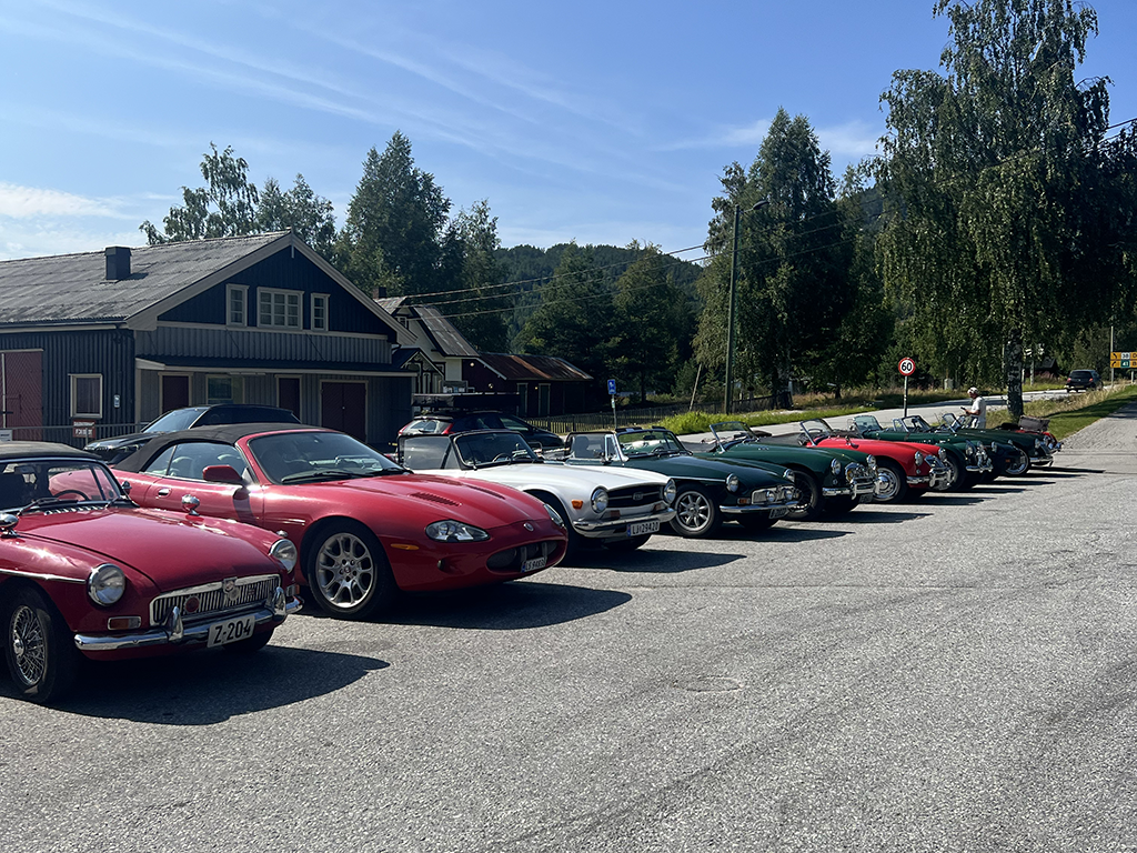 A line of classic and sports cars parked on a gravel lot beside a rural building, with trees and a hill in the background under a clear blue sky.