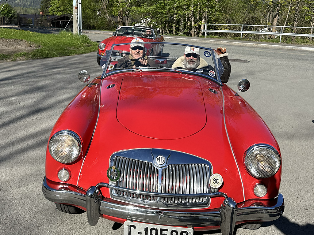Two older men driving a red vintage MG convertible car on a paved road, with a red sports car following behind. The men are wearing sunglasses and caps, and one is waving. The background has greenery and a white fence.