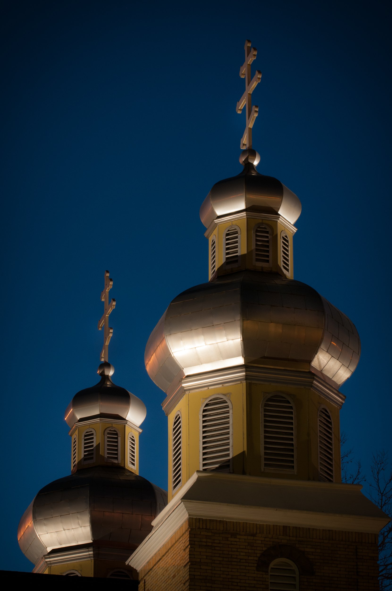 Nighttime view of the domed tops of a church with illuminated metallic surfaces and crosses on top, set against a dark blue sky.