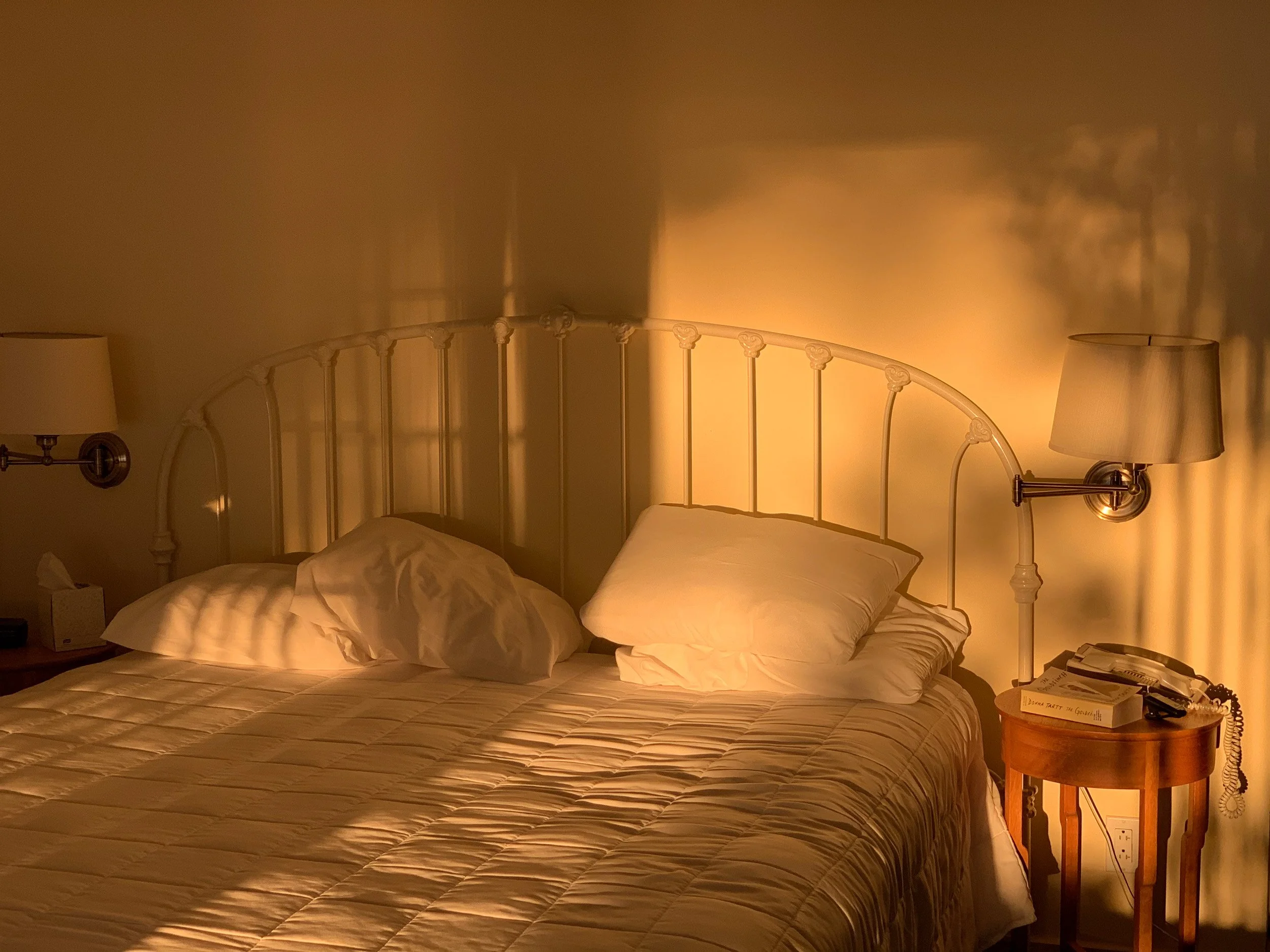 A cozy bedroom with a bed, pillows, and warm lighting from wall lamps. A nightstand holds books and a telephone.