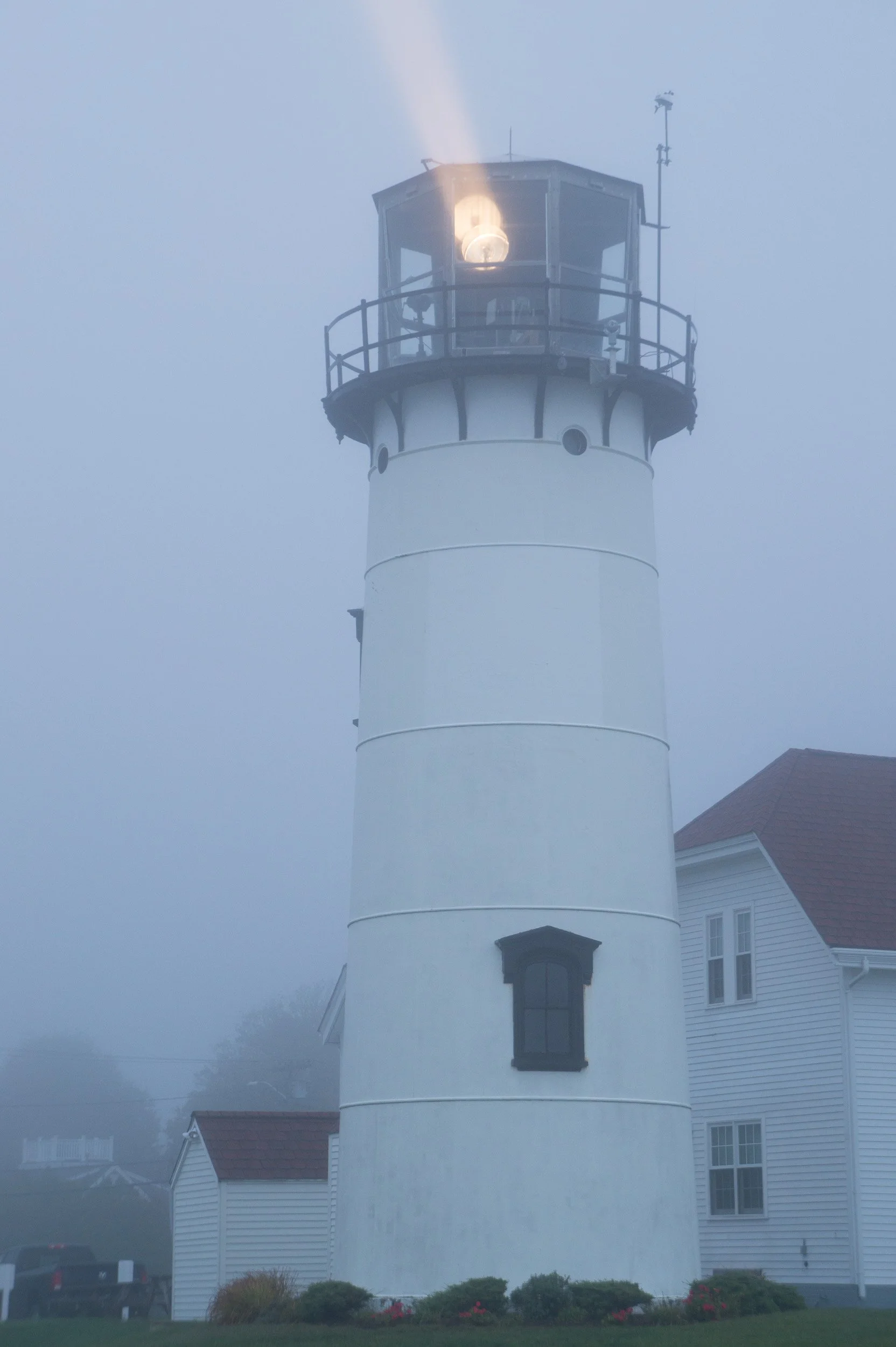 A white lighthouse with a black railing at the top, emitting a bright light through its lens in foggy conditions, adjacent to white houses with red roofs.