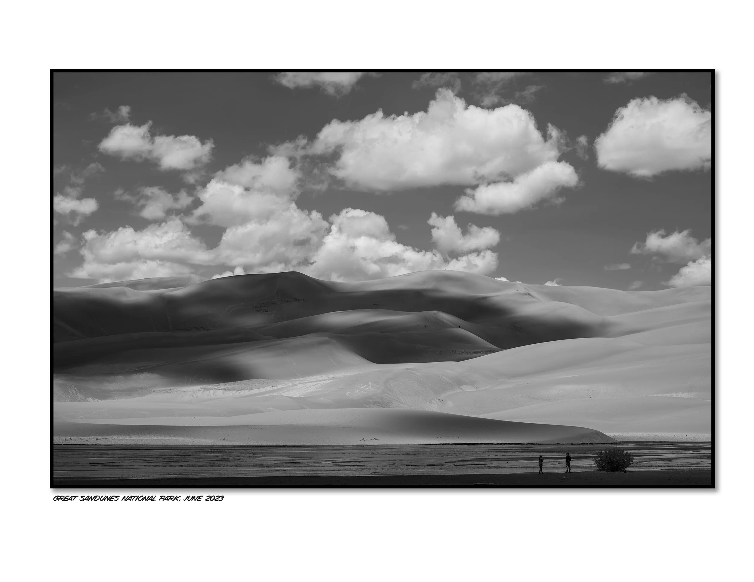 Black and white photo of sand dunes with two people standing on the ground near a bush, under a sky with scattered clouds. The caption reads "Great Sand Dunes National Park, June 2023."