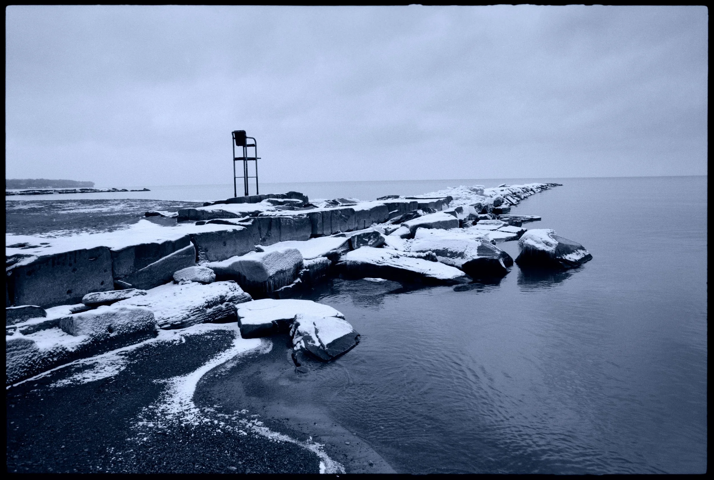 A rocky shoreline on a cloudy winter day with snow-covered rocks and calm water, featuring a metal lifeguard chair on a distant pier extending into the water.