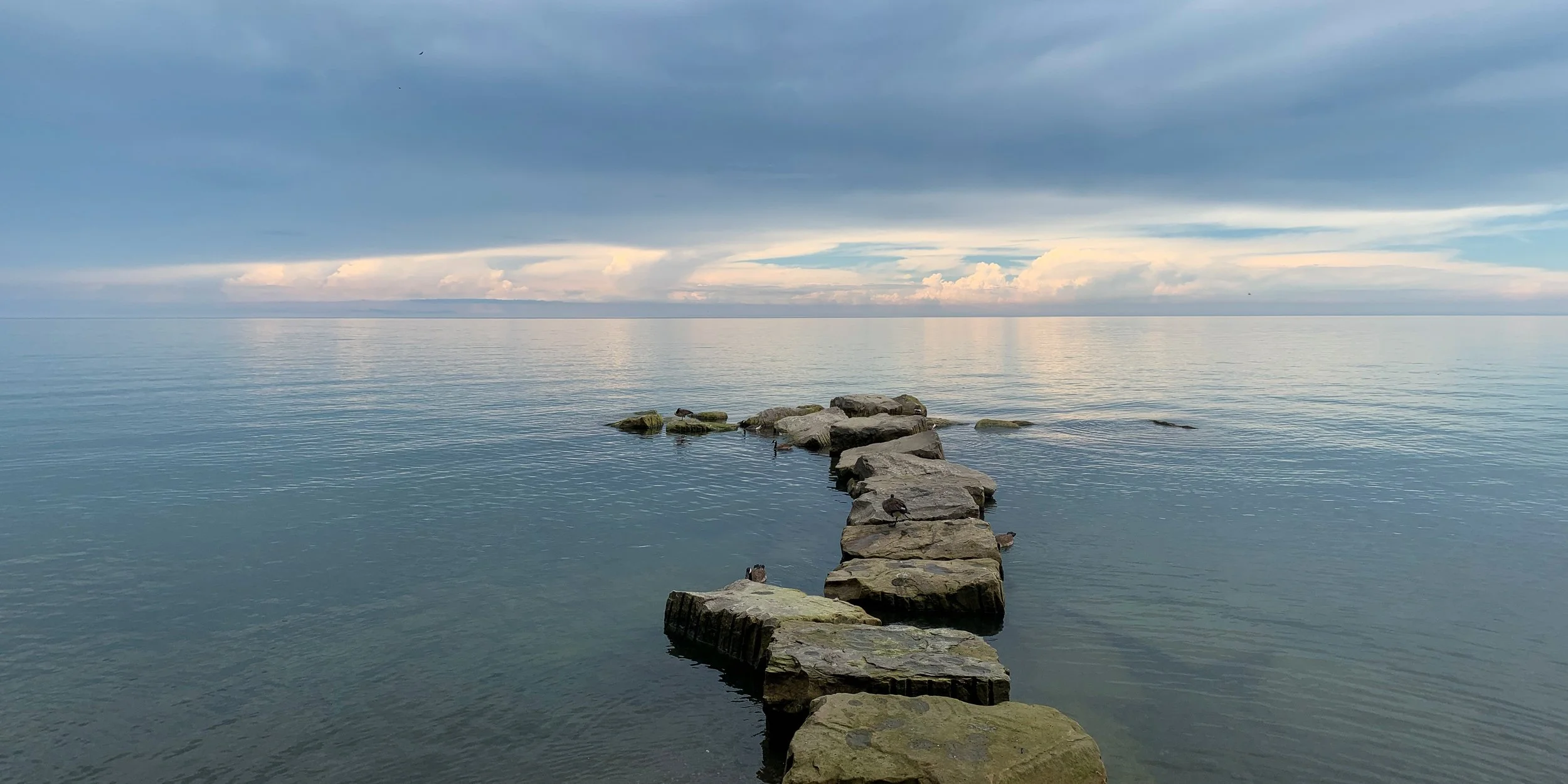 A stone pier extending into a calm body of water under a cloudy sky with patches of blue and sunlight.