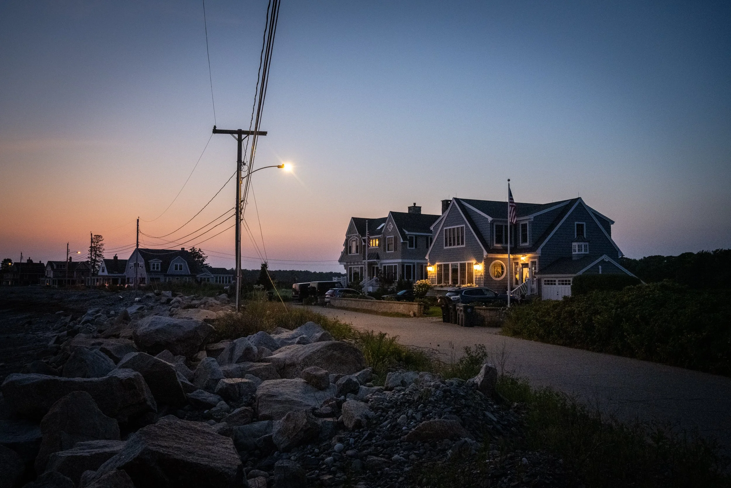 A street scene during sunset featuring a large house with lit windows, a flagpole with an American flag, power lines with a street lamp, SUVs parked in front, rocks along the roadside, and a backdrop of smaller houses and trees.