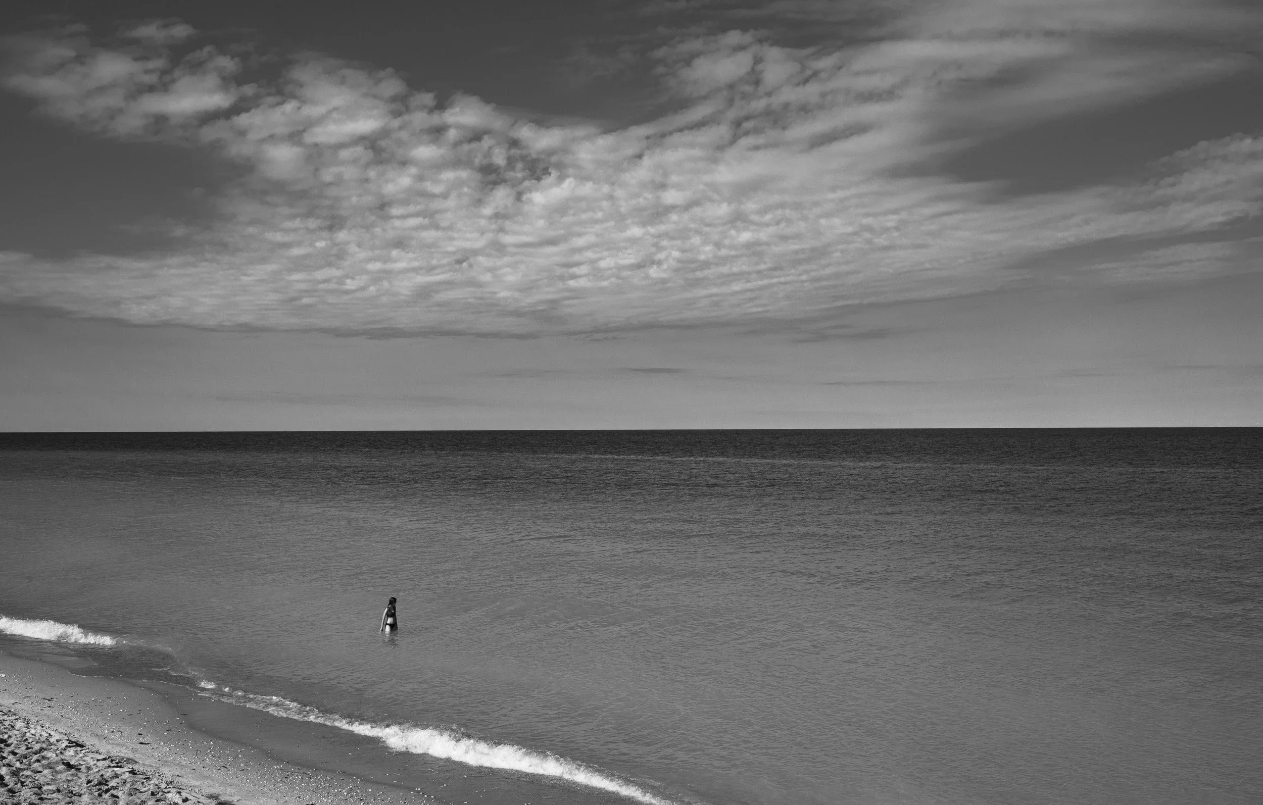A person standing in the water at the beach with waves on the shore, under a partly cloudy sky.