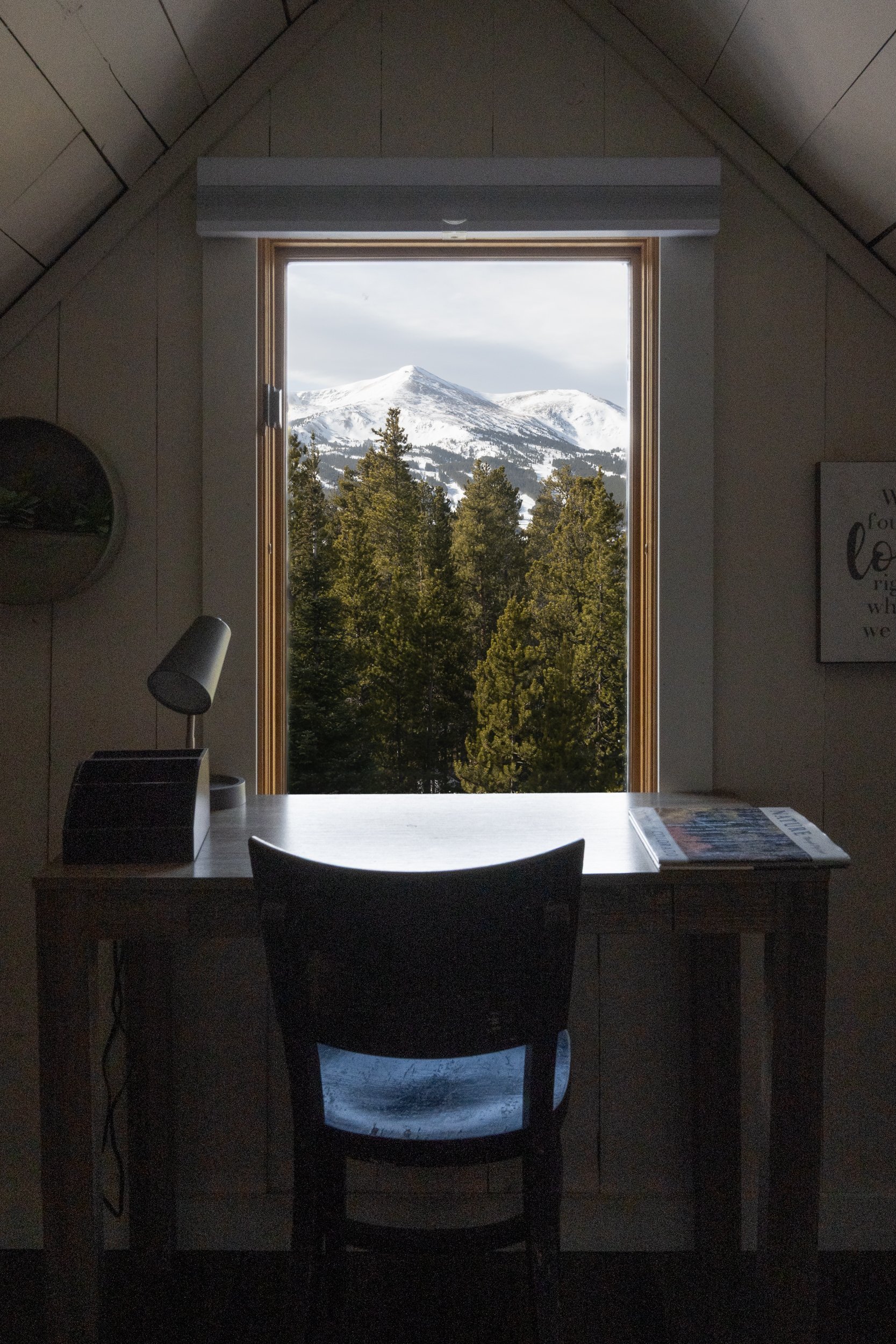 Interior view of a cozy room with a wooden desk and chair positioned in front of a window with a mountain and snow-covered trees outside.