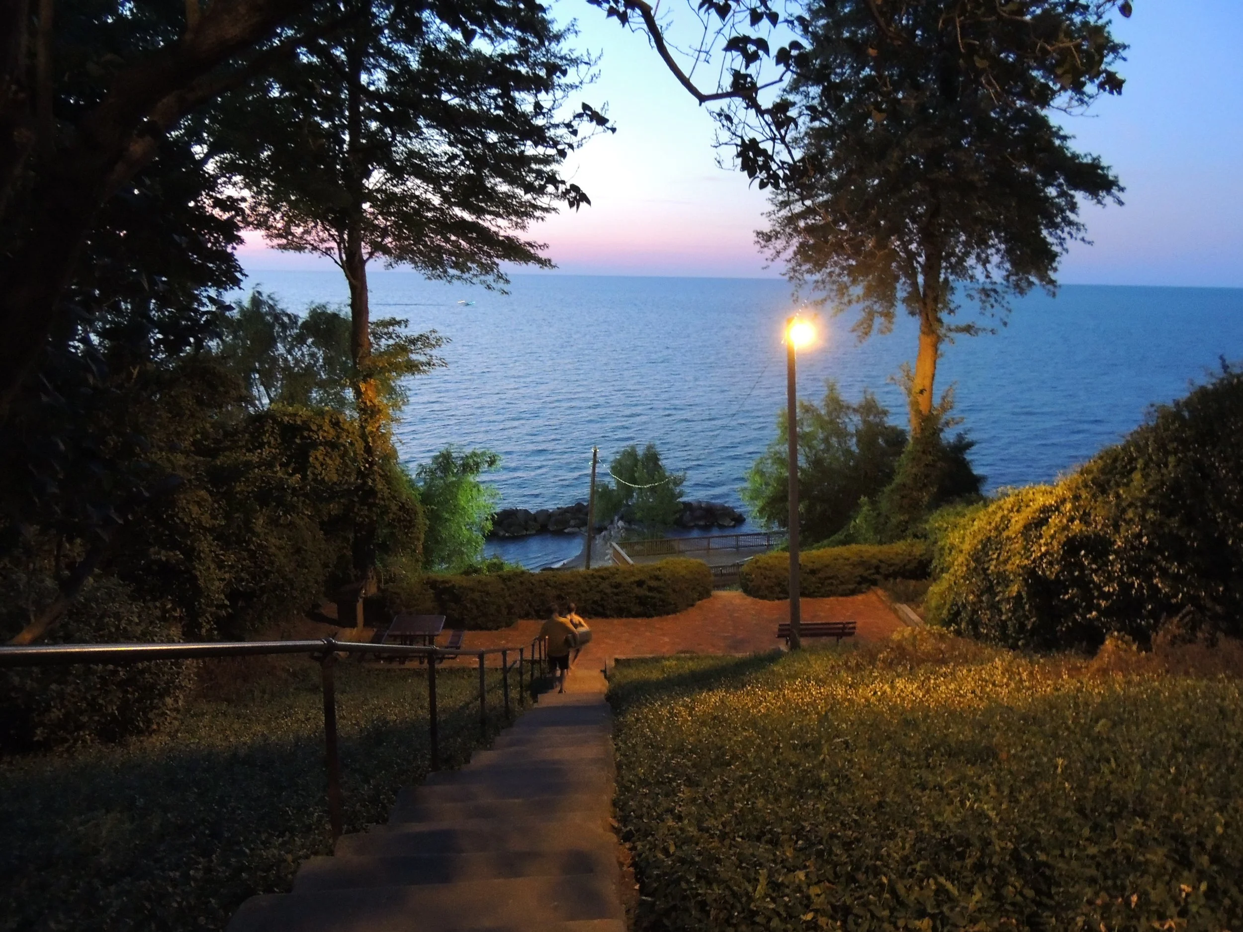 A staircase leads down to a lakeside park area illuminated by a streetlamp, with trees and bushes surrounding the scene, and a calm lake in the background during dusk.