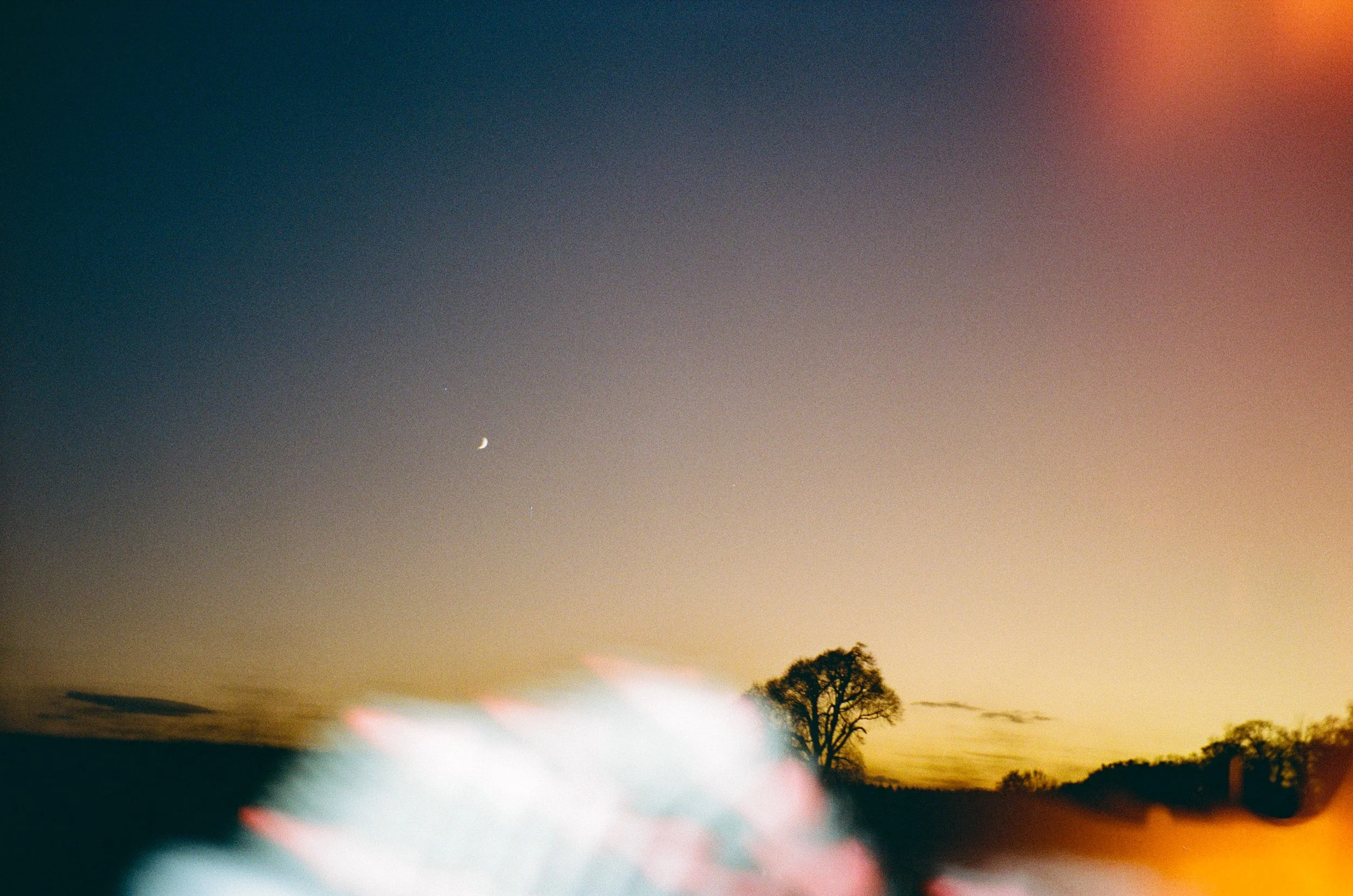 A twilight sky with a crescent moon and a bright star, silhouettes of trees on the horizon, and some blurred objects in the foreground.