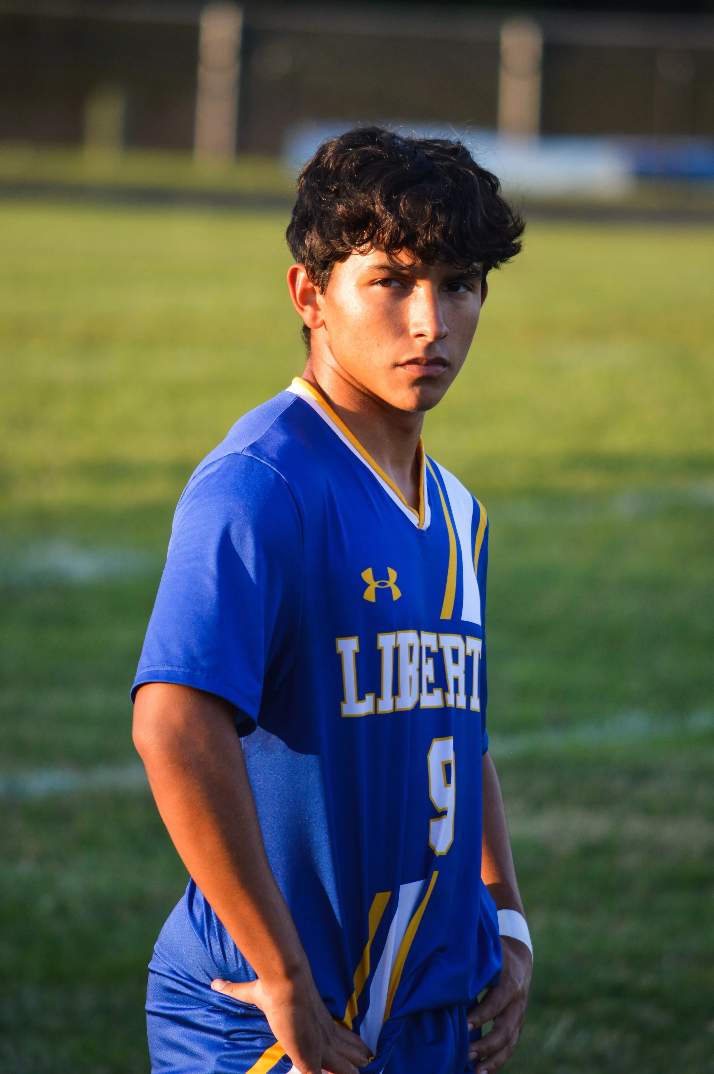 A young male athlete wearing a blue soccer jersey with yellow and white accents and the number 9, standing on a grass field during sunset.